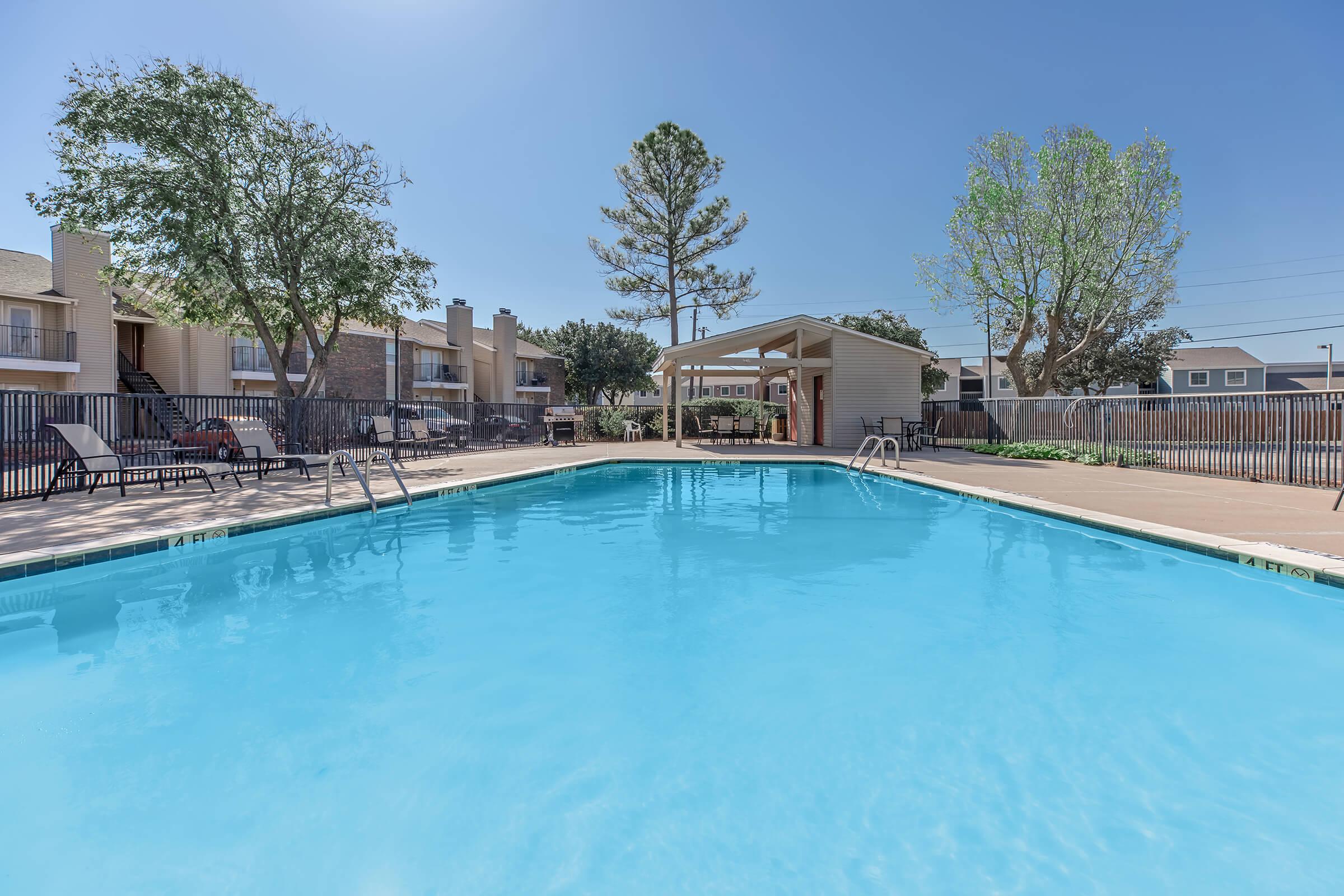 A clear blue swimming pool surrounded by concrete decking, with lounge chairs on one side. In the background, there are residential buildings and trees under a bright blue sky. A shaded gazebo structure is also visible near the pool area, providing a relaxing spot.