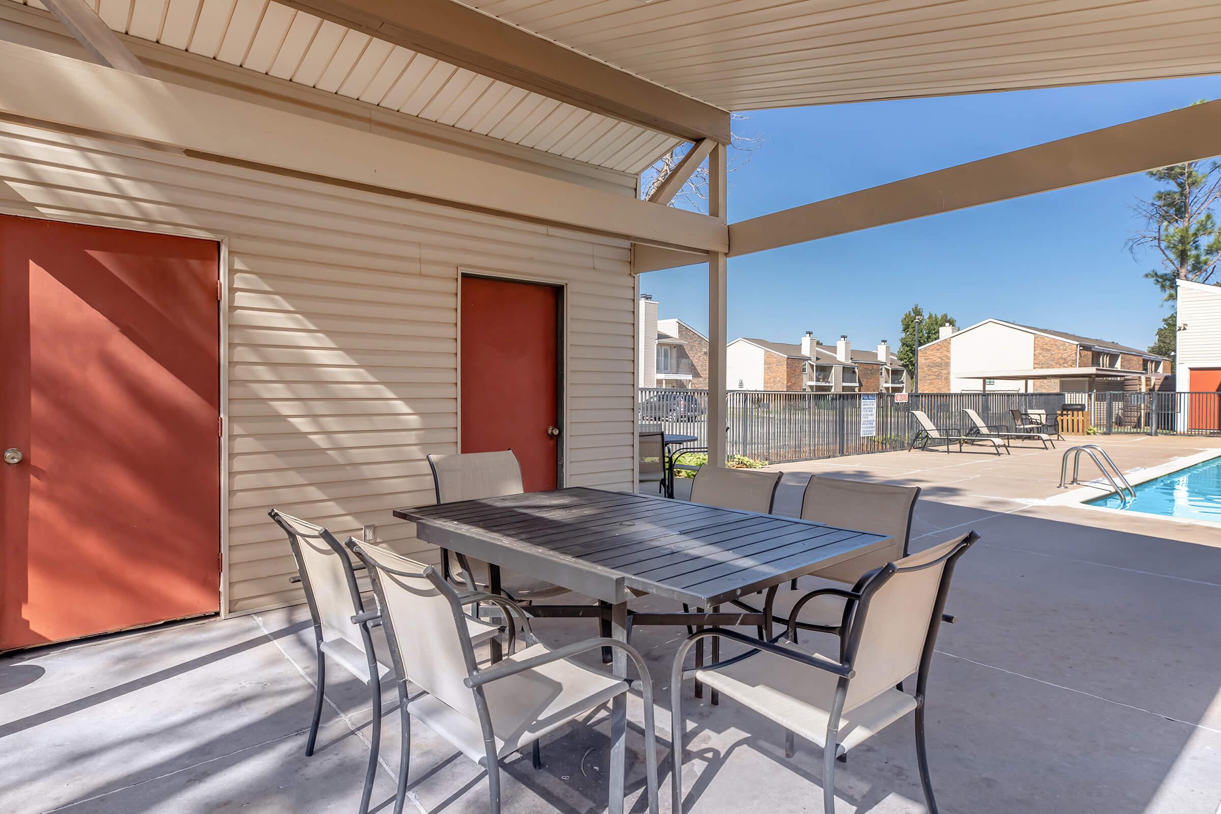 Outdoor lounge area featuring a metal table surrounded by six chairs, shaded by a roof. Two red doors lead to nearby rooms, and a swimming pool is visible in the background along with lounge chairs and well-maintained landscaping under a clear blue sky.