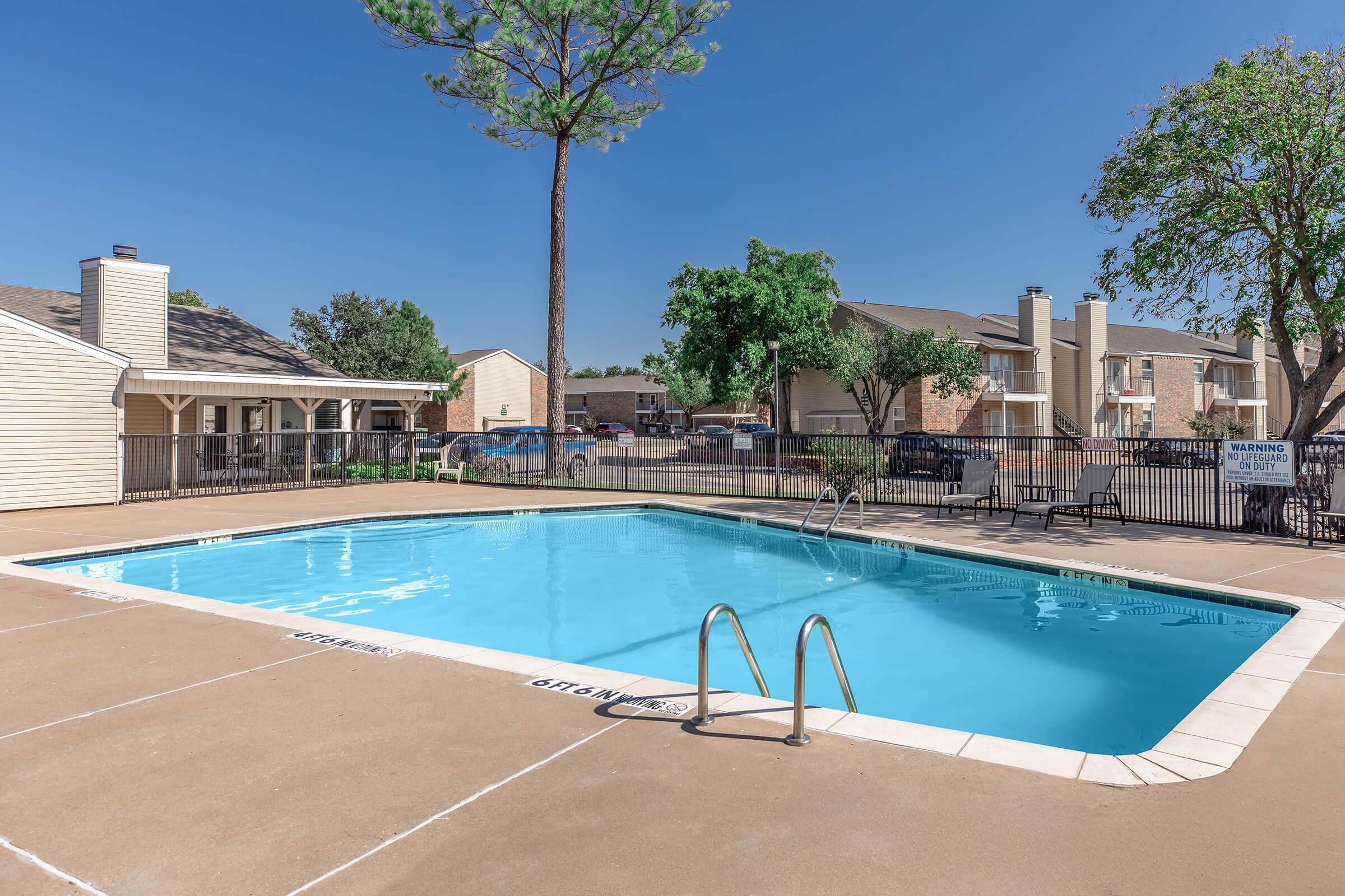 A clear blue swimming pool surrounded by a paved area and lounge chairs, with a few trees and buildings in the background. The sky is bright and sunny, creating a inviting atmosphere for relaxation and leisure. A sign is visible near the pool area.