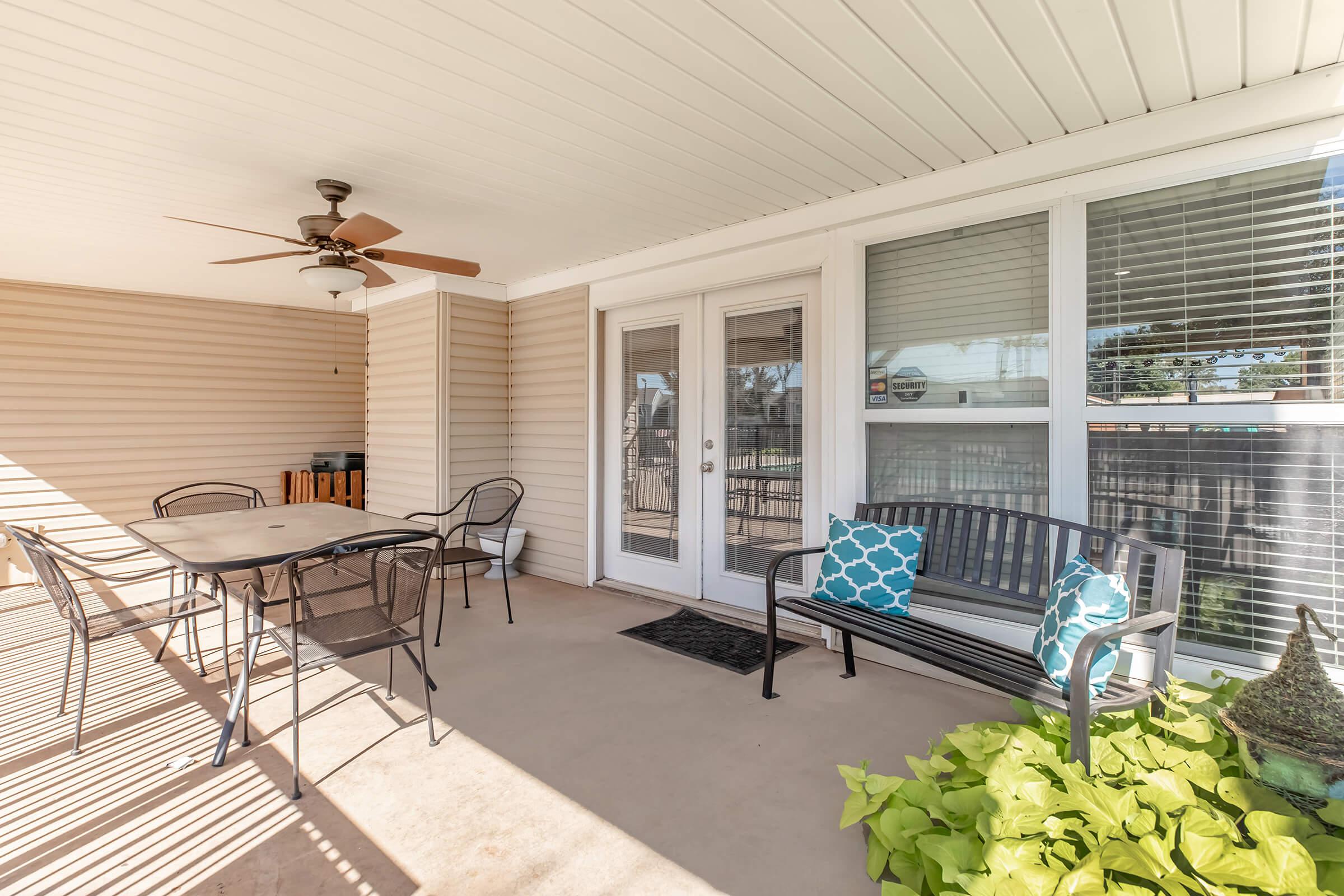 A covered patio area featuring a ceiling fan, a table with four chairs, a bench with decorative pillows, and potted plants. Large windows provide natural light, and the space is designed for relaxation and outdoor dining.