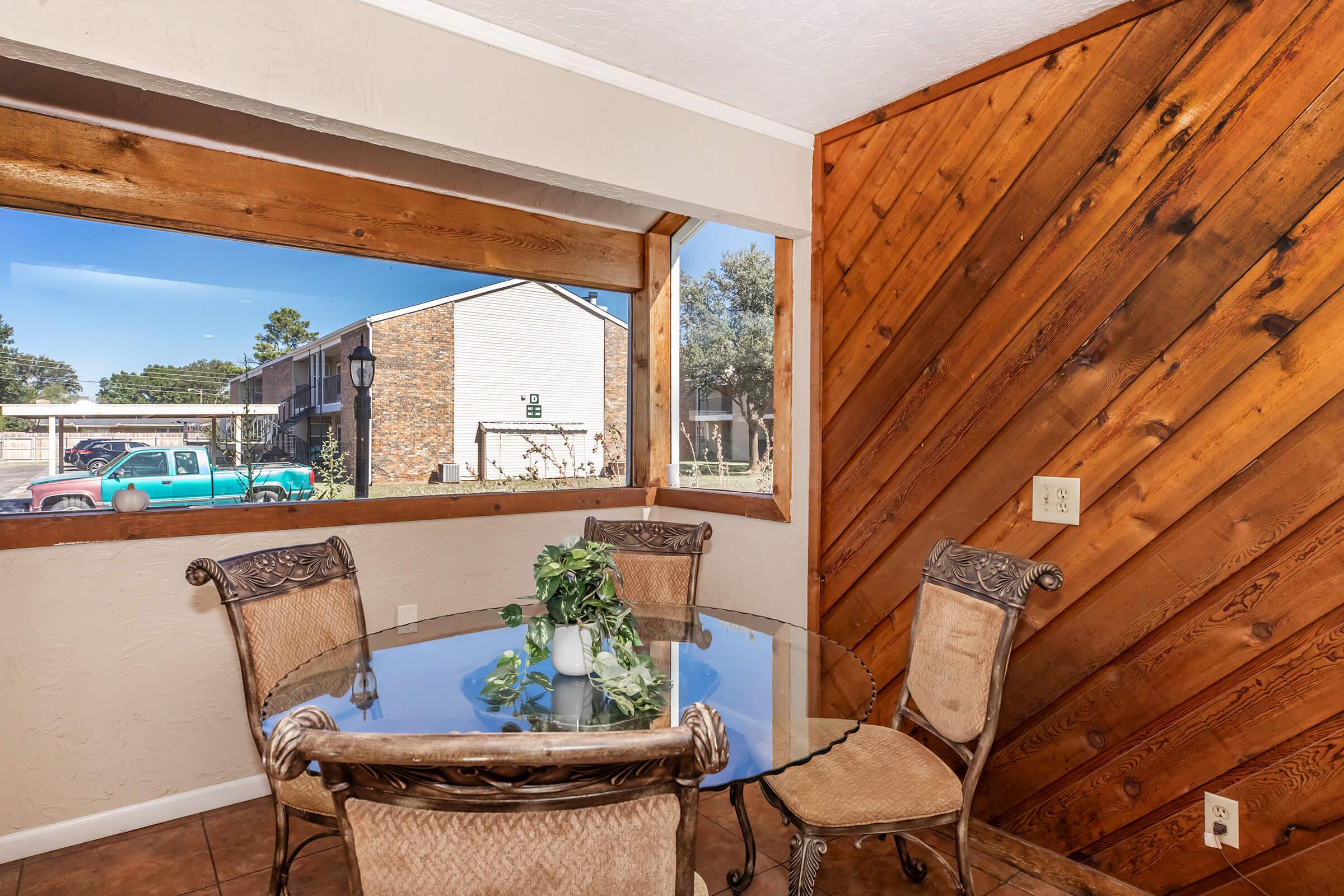 A cozy dining area featuring a circular glass table surrounded by four ornate chairs. The wooden walls add warmth to the space, and a large window offers a view of the outdoors, showcasing a parking area with a vintage green truck. A small plant centerpiece enhances the inviting atmosphere.