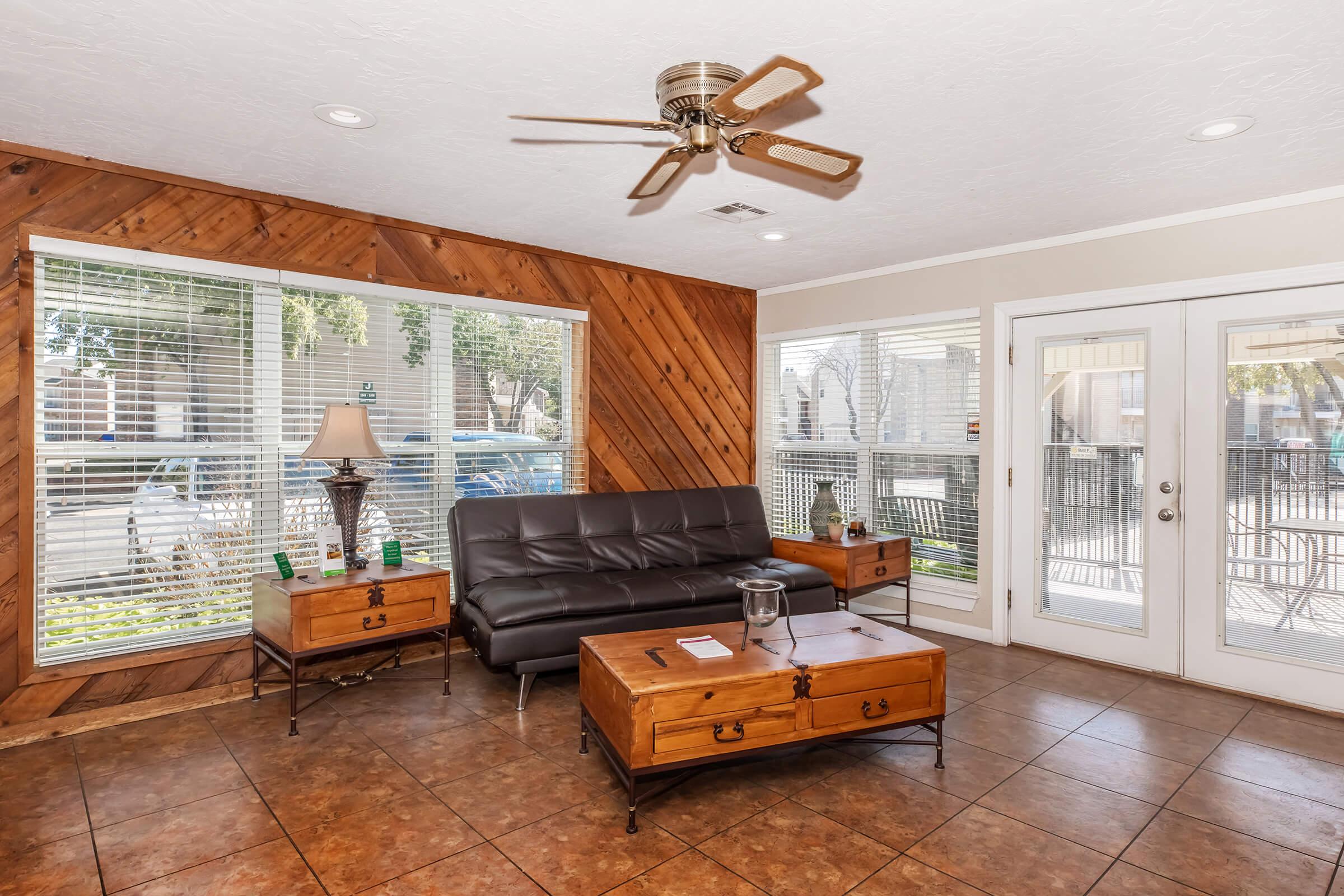 A modern living room featuring wooden accent walls, a brown leather sofa, and two wooden coffee tables. A lamp sits on one of the tables, and large windows provide natural light and a view outside. A ceiling fan hangs above, enhancing the cozy atmosphere.