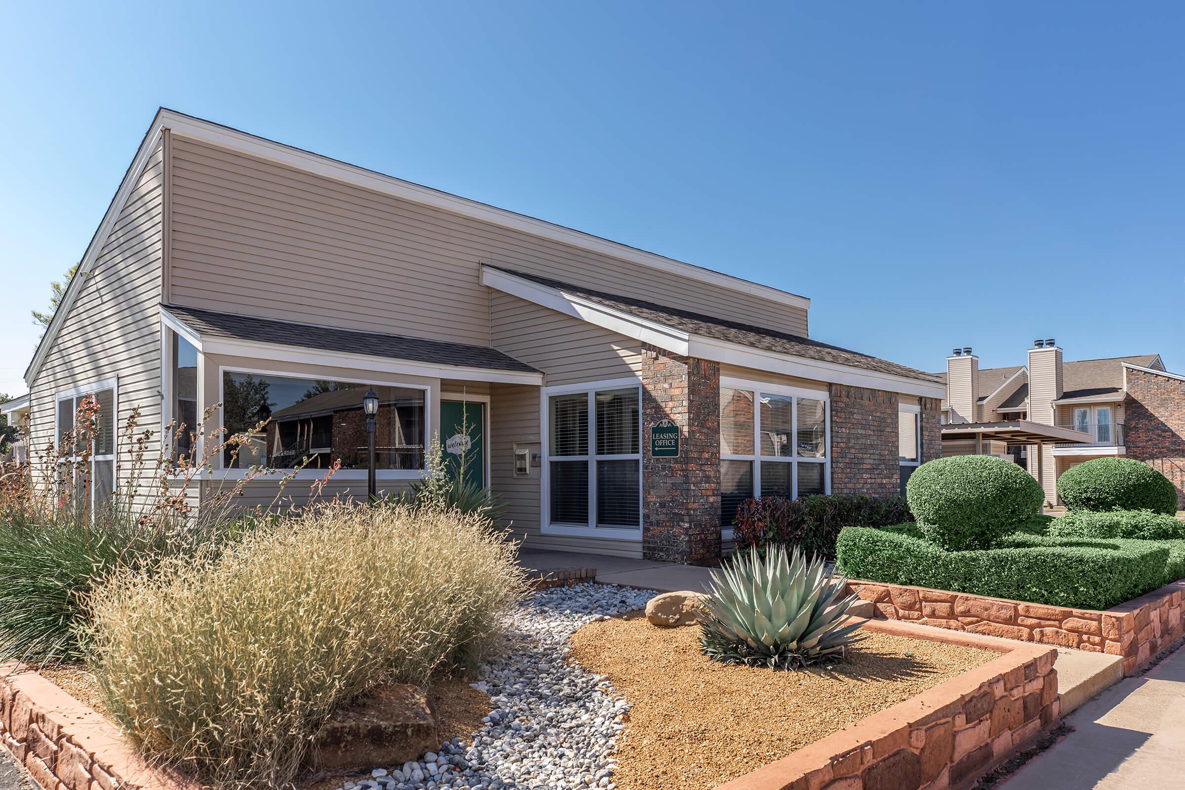 A modern single-story house with a combination of siding and stone accents. The front yard features decorative gravel, succulents, and carefully shaped shrubs. Clear blue skies above add to the sunny atmosphere. The house has a welcoming porch area, enhancing its charming curb appeal.