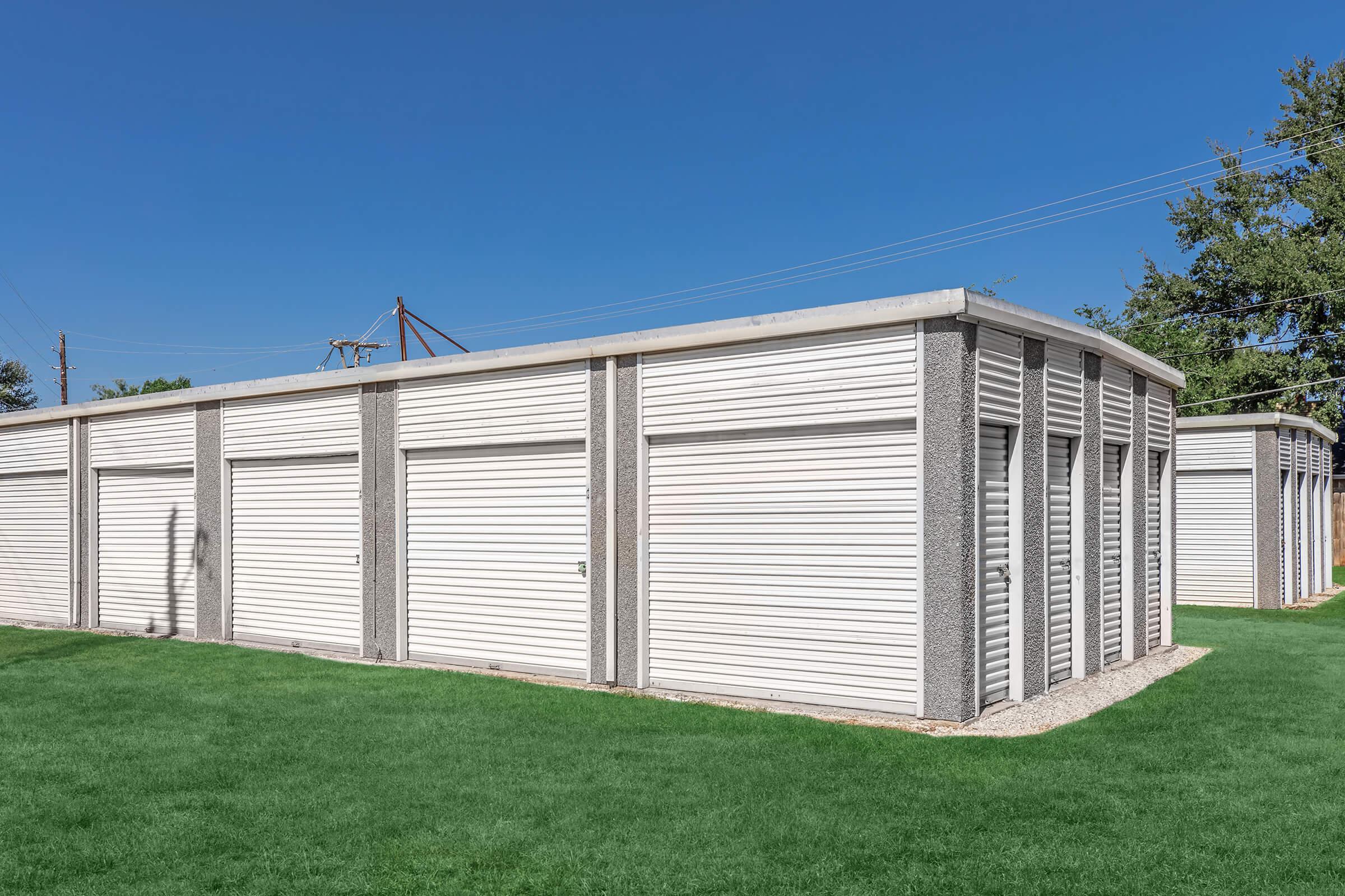 Storage units with roll-up doors arranged in a row, set against a clear blue sky. The units are situated on green grass, and there are power lines in the background. The overall scene is neat and organized, showcasing a functional storage facility.