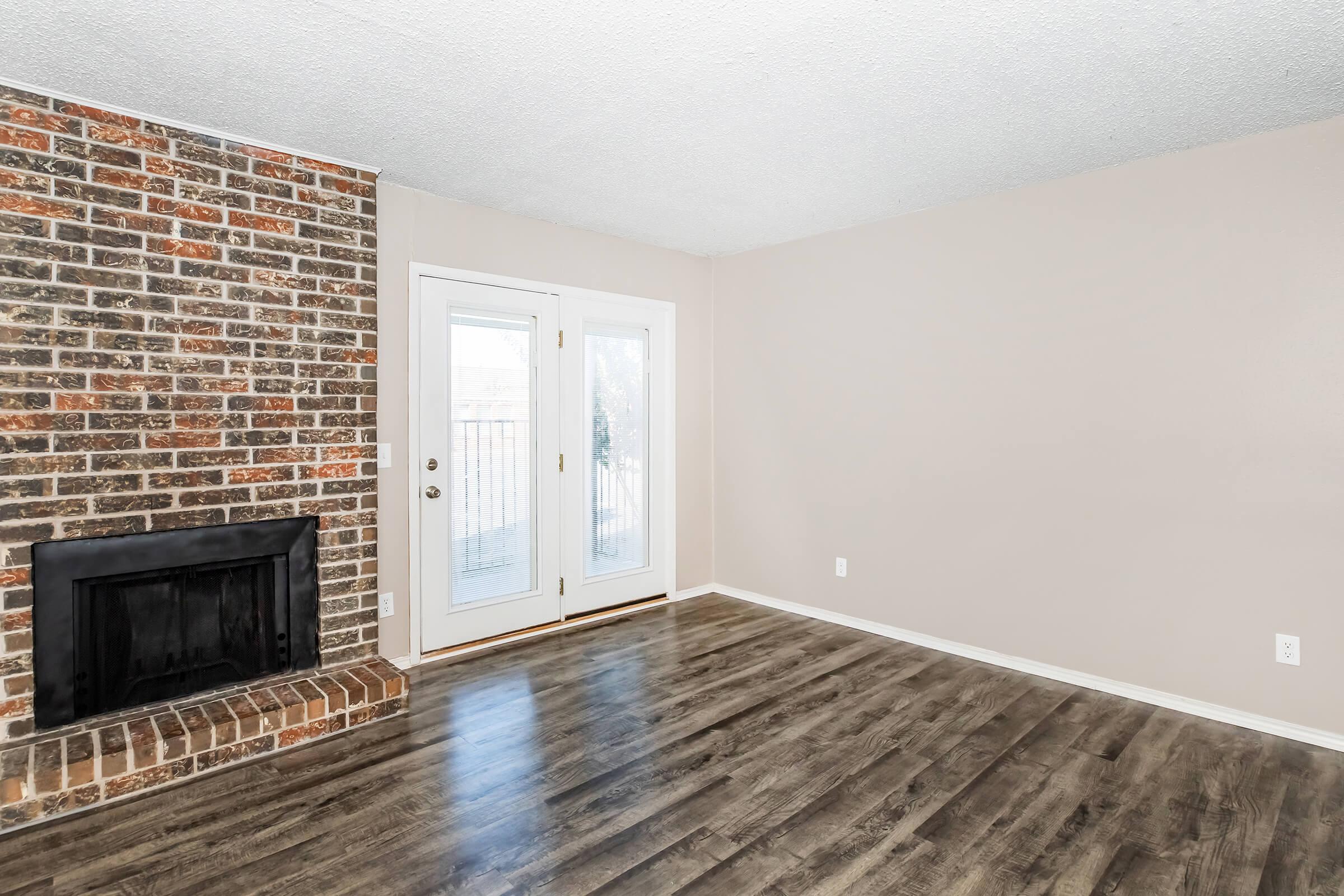 Spacious living room with a brick fireplace on the left, light-colored walls, and hardwood flooring. Double glass doors lead to an outdoor space. The room is well-lit, featuring a minimalist design and ample open space for furniture arrangement.