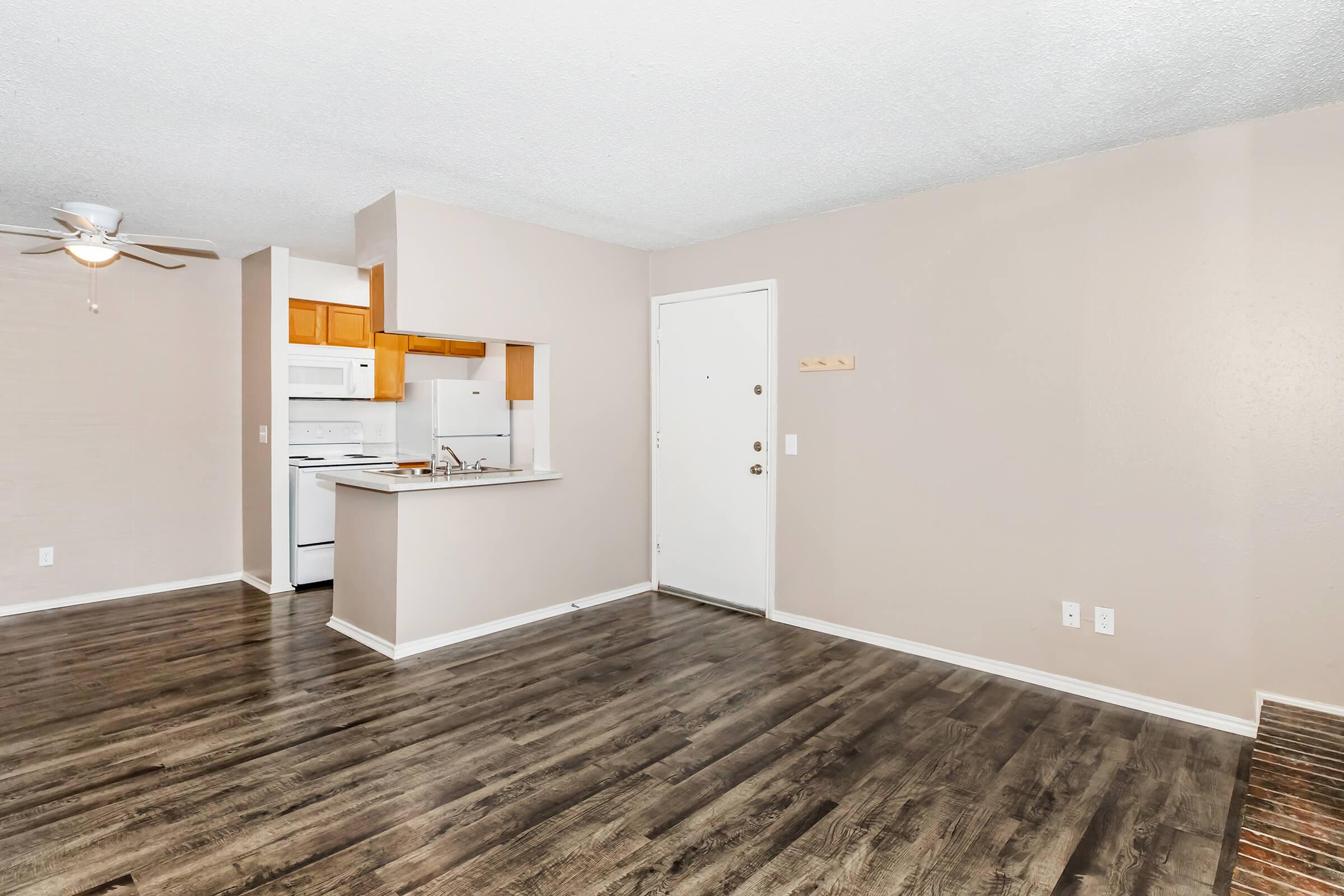 A spacious living area featuring light-colored walls and faux wood flooring. A ceiling fan is visible, along with an open kitchen area that has wooden cabinets and white appliances. The entrance door is located on one side, enhancing the room's inviting ambiance.