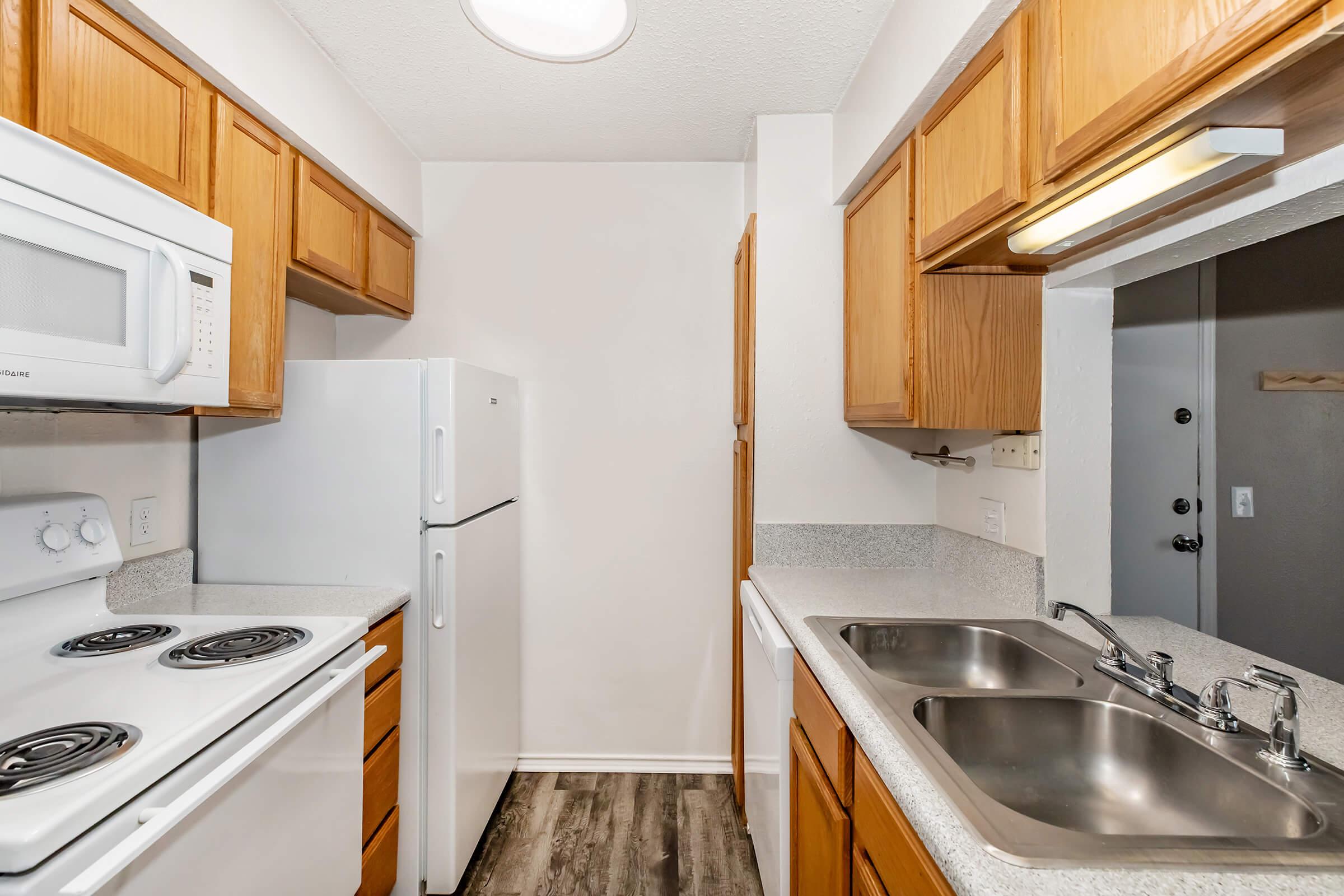 A small kitchen featuring light wood cabinets, a white refrigerator, a microwave above the stove, and a double sink. The countertop is gray, and the flooring is textured. There is a door that leads outside visible in the background.