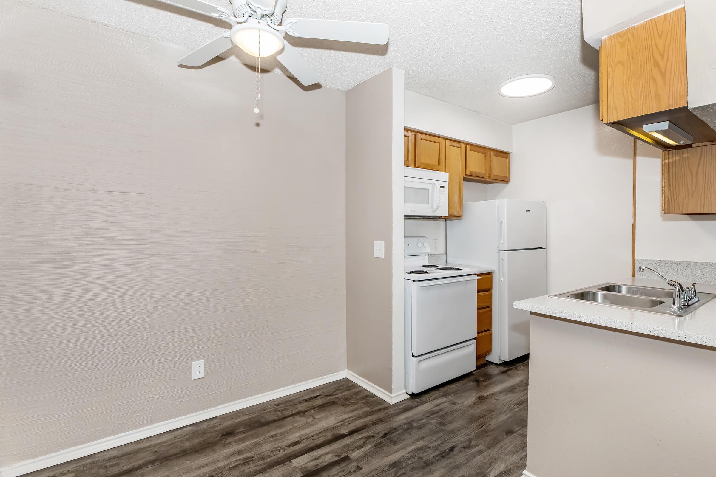 A small kitchen space featuring light-colored walls, a ceiling fan, wooden cabinets, a white stove, and a refrigerator. There is a sink with a countertop, and a laminate floor that adds warmth to the area. Natural light comes from a ceiling fixture, creating a bright and inviting atmosphere.