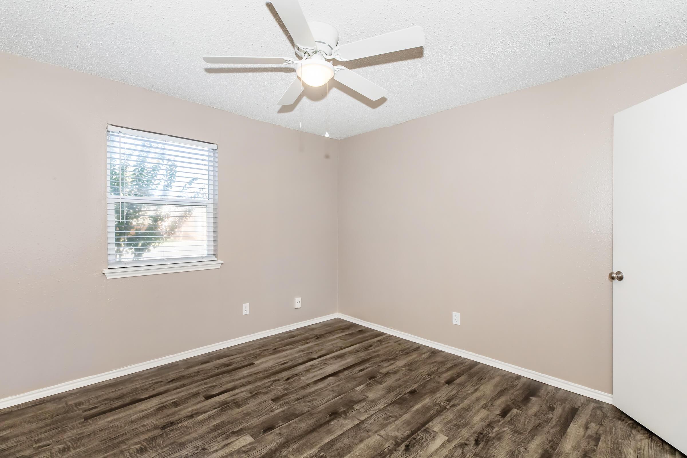 A simple, empty bedroom with light brown walls and a ceiling fan. The room features a window with blinds allowing natural light in, a light-colored door, and dark wood-style flooring. There are no furnishings, creating a spacious and minimalistic feel.