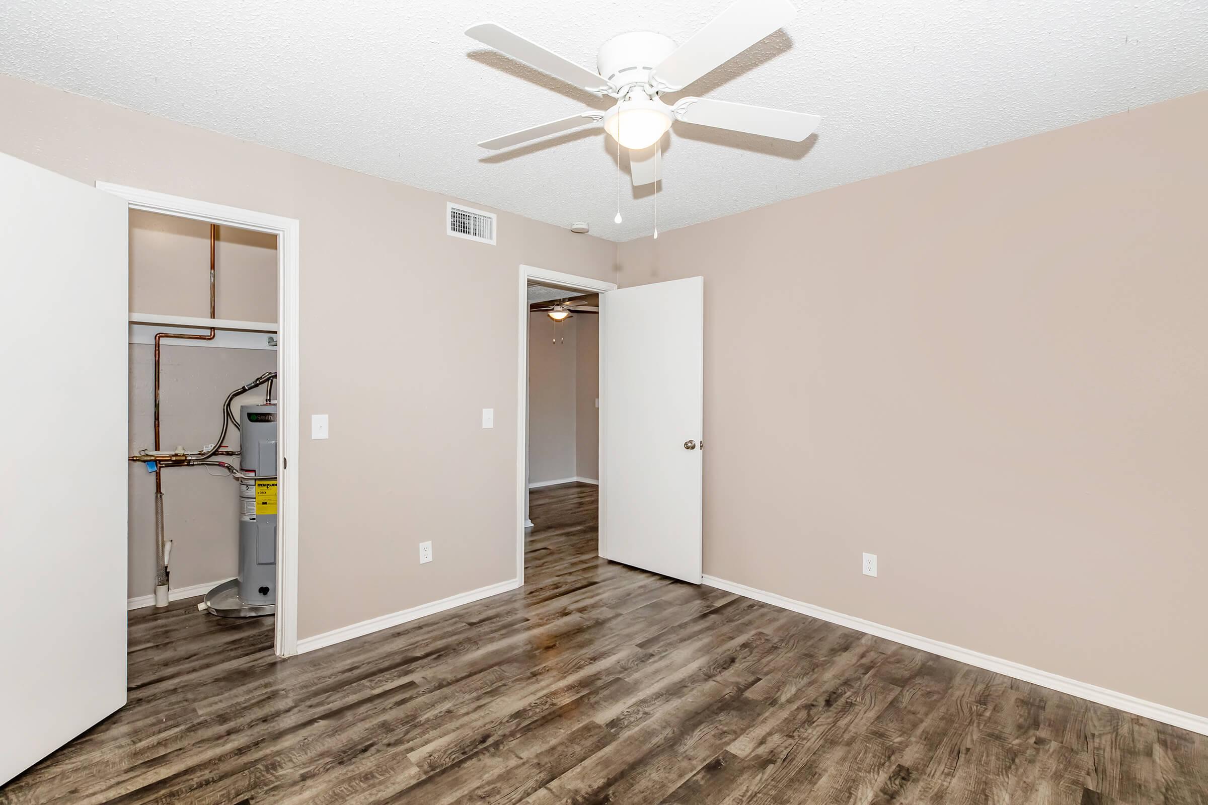 A well-lit room with beige walls and dark wooden flooring. It features a ceiling fan, a closet on the left with plumbing visible, and an open door leading to another room in the background. The overall appearance is clean and modern, suitable for various uses.