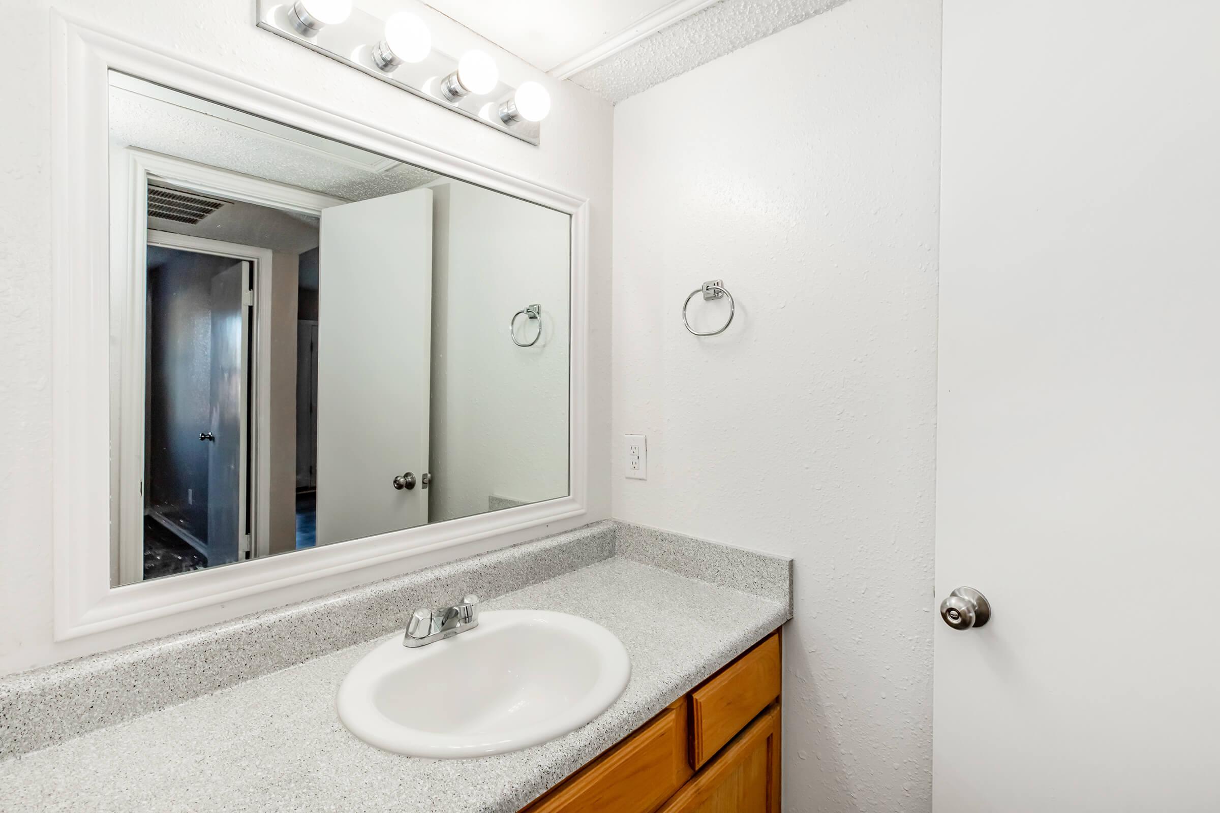 A well-lit bathroom featuring a white wall-mounted mirror above a light gray countertop with a sink. The wooden cabinet below the sink complements the space. A towel rack is mounted near the mirror, and a partially open door leads to another area. The walls are painted white, enhancing the airy feel.