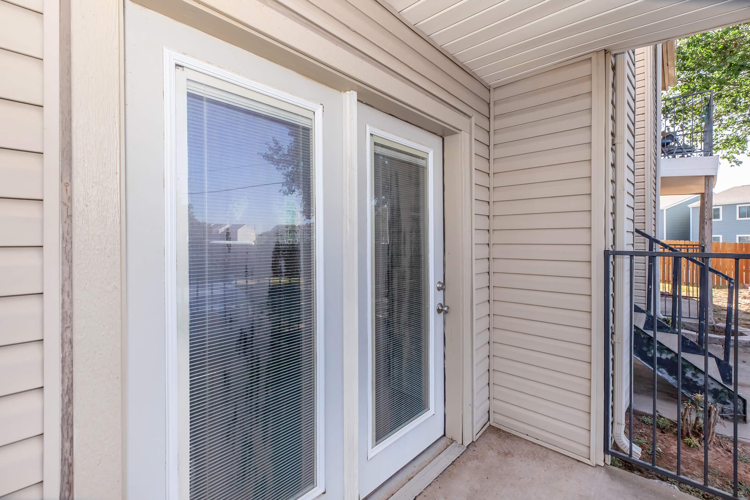 Double glass doors leading to a porch area, framed by light-colored walls. The doors feature a textured pattern and a small handle, with a glimpse of the outdoors visible through the glass. To the right, there's a short staircase and a railing, surrounded by a neutral-toned exterior.