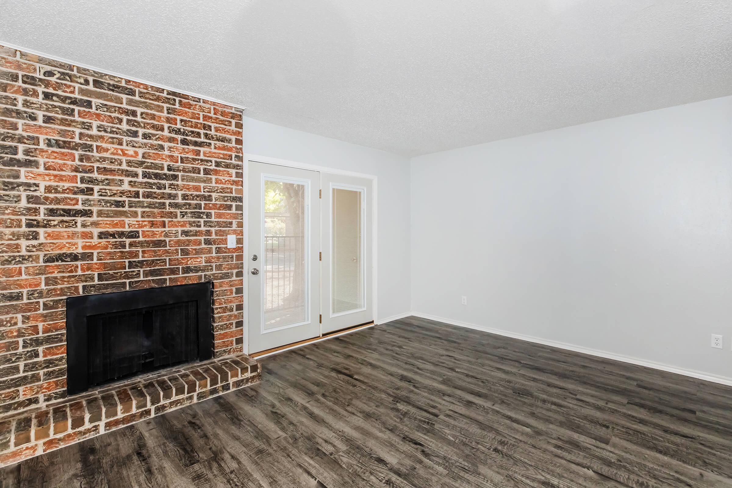 A spacious living room featuring a brick accent wall with a fireplace, light-colored walls, and a large window with double doors leading outside. The flooring is dark wood, creating a warm and inviting atmosphere.