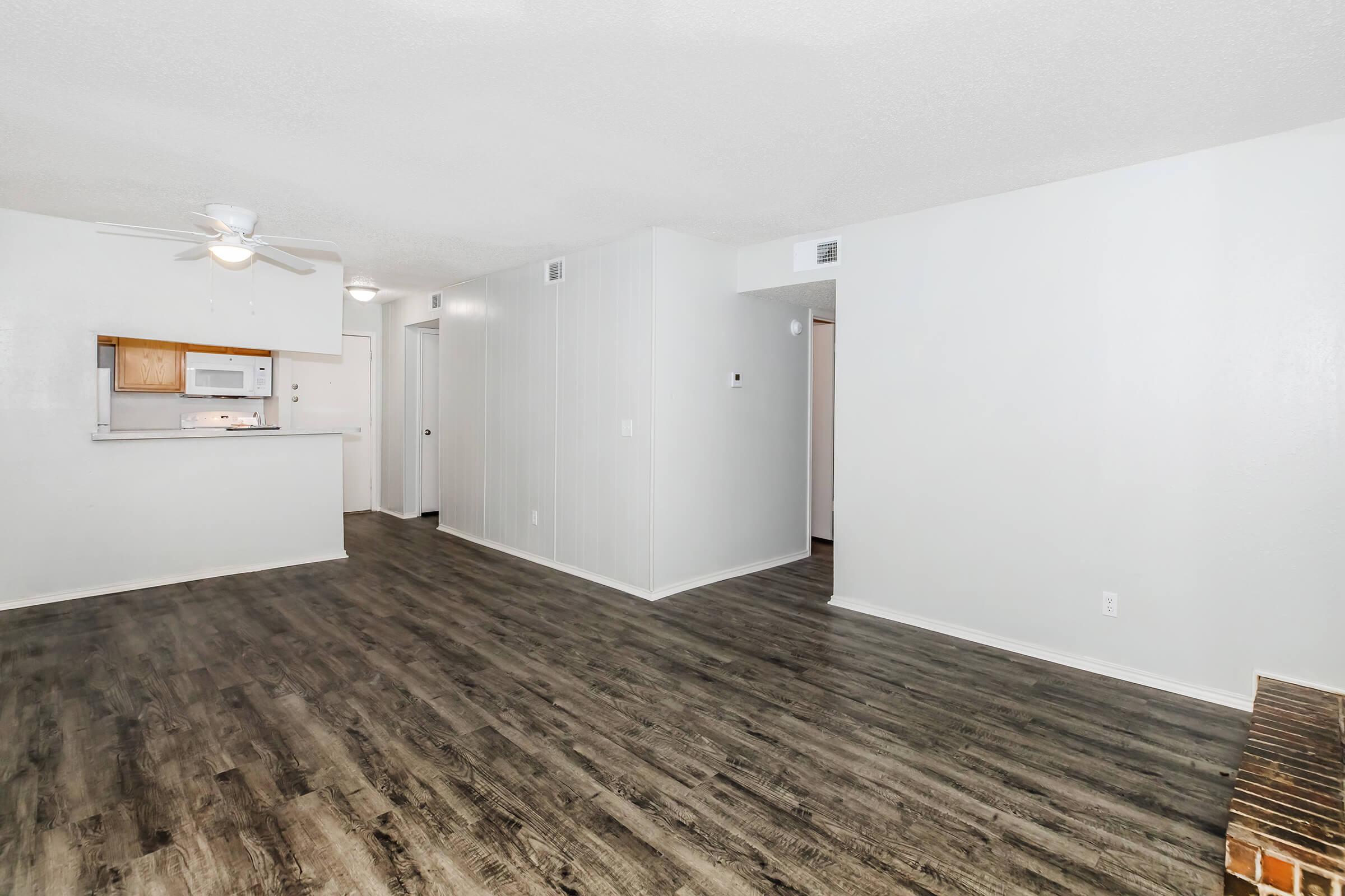 An open-concept living area featuring light-colored walls and a modern wood-like floor. A ceiling fan is visible, and the space leads to a small kitchen with a microwave. The overall design is simple and inviting, with ample room for furniture arrangement.