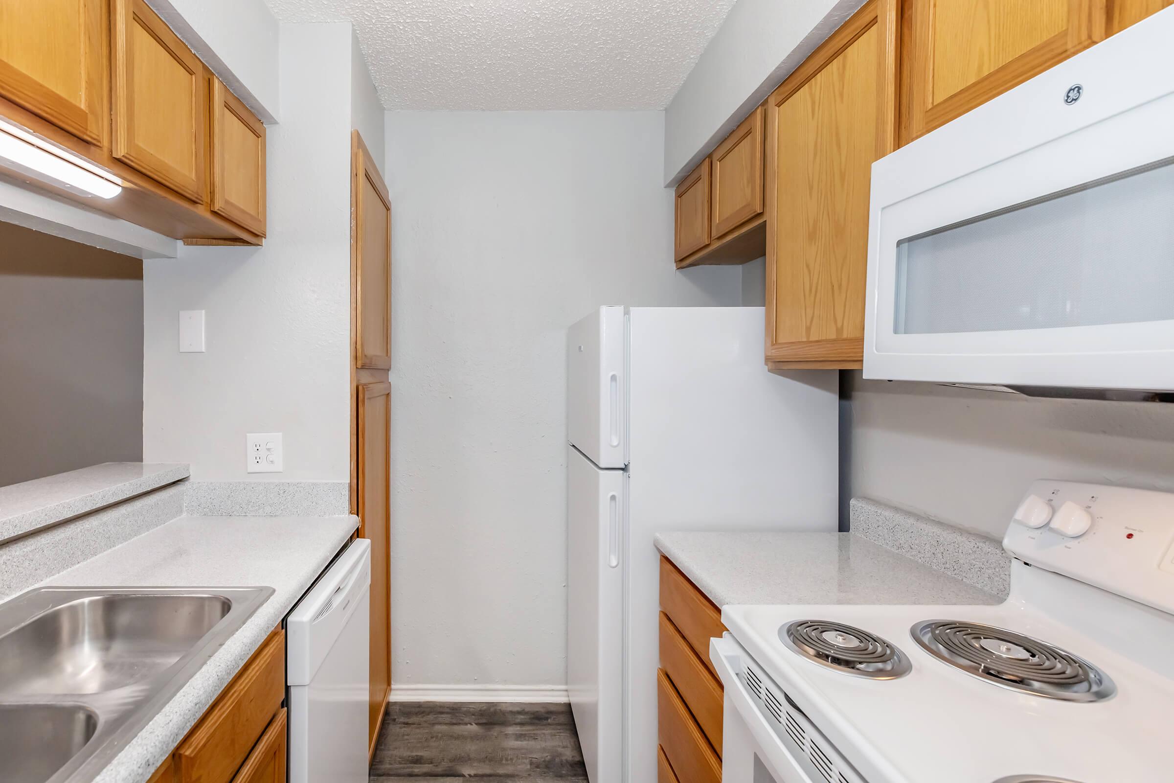 A modern kitchen featuring light wood cabinetry, white appliances including a refrigerator, stove, and microwave, along with a double sink. The countertops are gray, and the walls are painted a light color, creating a clean and spacious look. The flooring is dark, contrasting with the cabinetry.