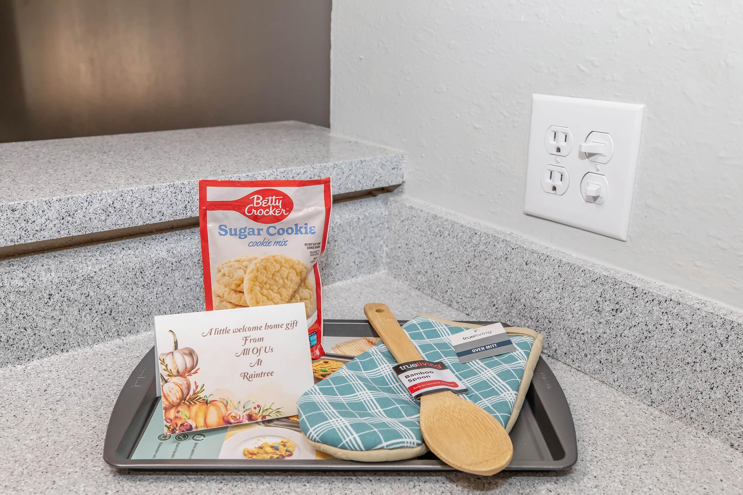 A countertop scene featuring a packet of Betty Crocker Sugar Cookie mix, a greeting card with a fall theme, a baking sheet, a wooden spoon, and a blue and white oven mitt, all arranged neatly. Electrical outlets are visible in the background.