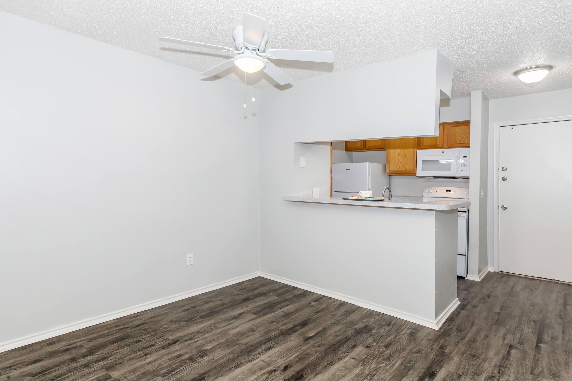 A contemporary apartment interior featuring a light-colored wall, a ceiling fan, and laminate flooring. There's a small kitchen area with wooden cabinets, a microwave, and a stove, visible from an open living space. A door leading outside is in the background.
