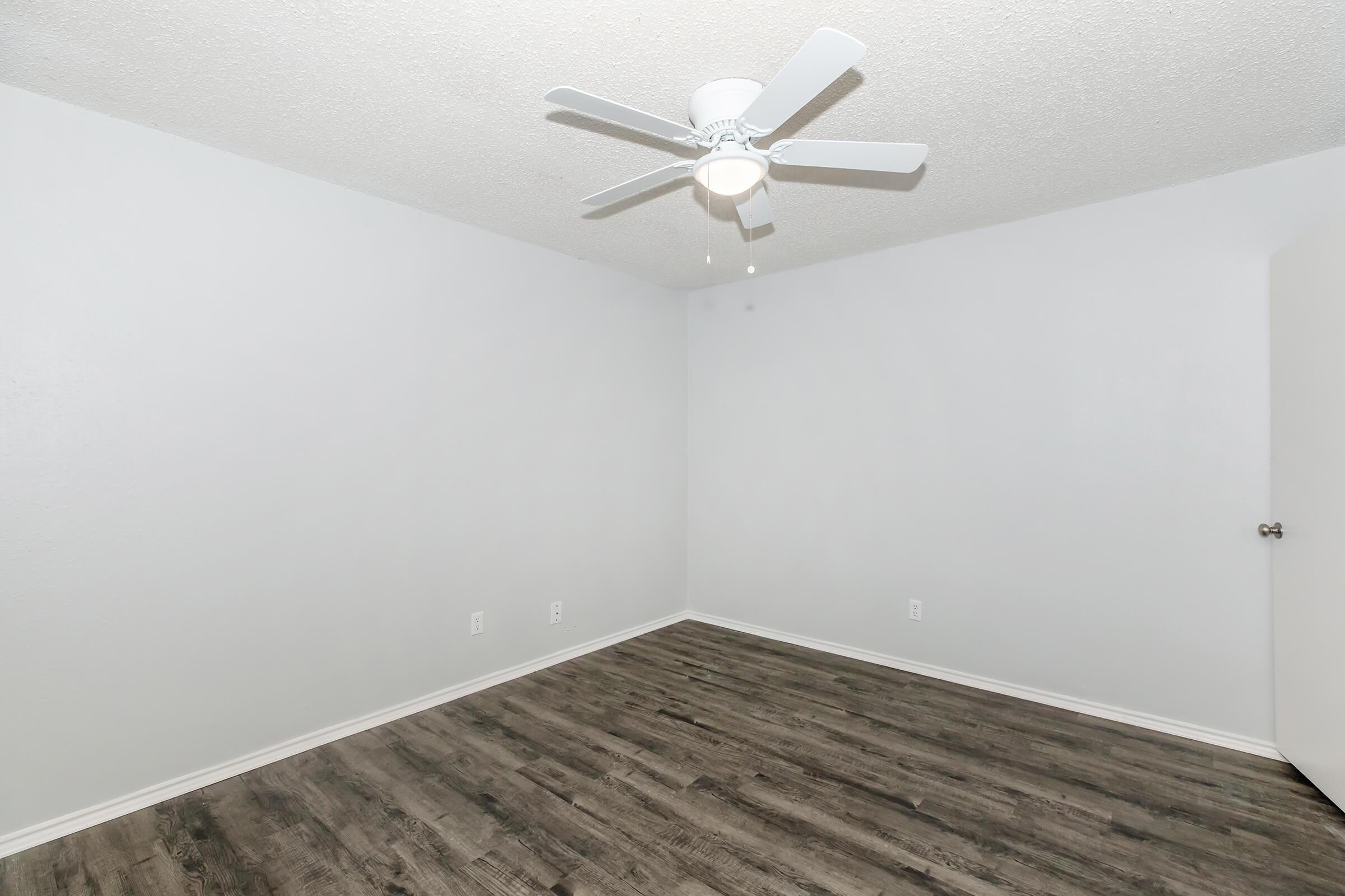 Empty room with light gray walls and dark wood-like flooring. A white ceiling fan is mounted in the center, providing light. The room has a closed door on one side and no furniture or decorations, creating a spacious and minimalistic atmosphere.