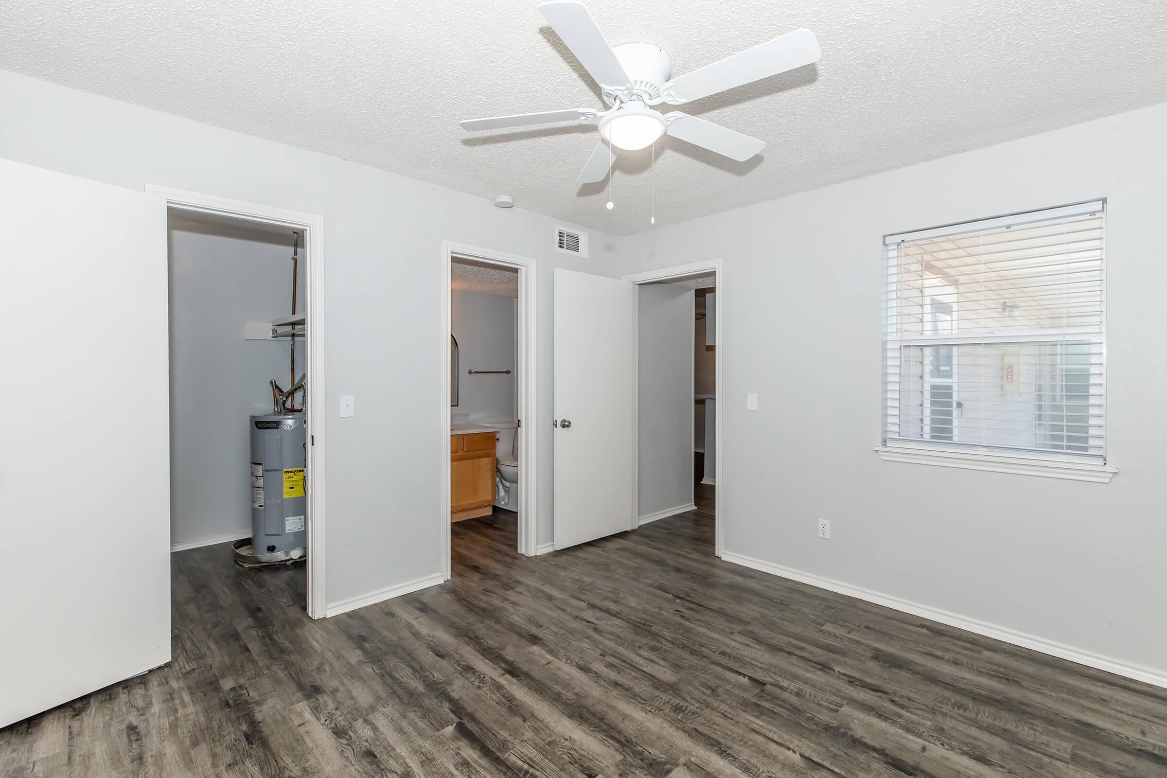 A well-lit, neutral-colored room with a ceiling fan. There are three doorways leading to other areas, including a bathroom and laundry space, and a window allowing natural light. The floor features a modern wood-like finish.