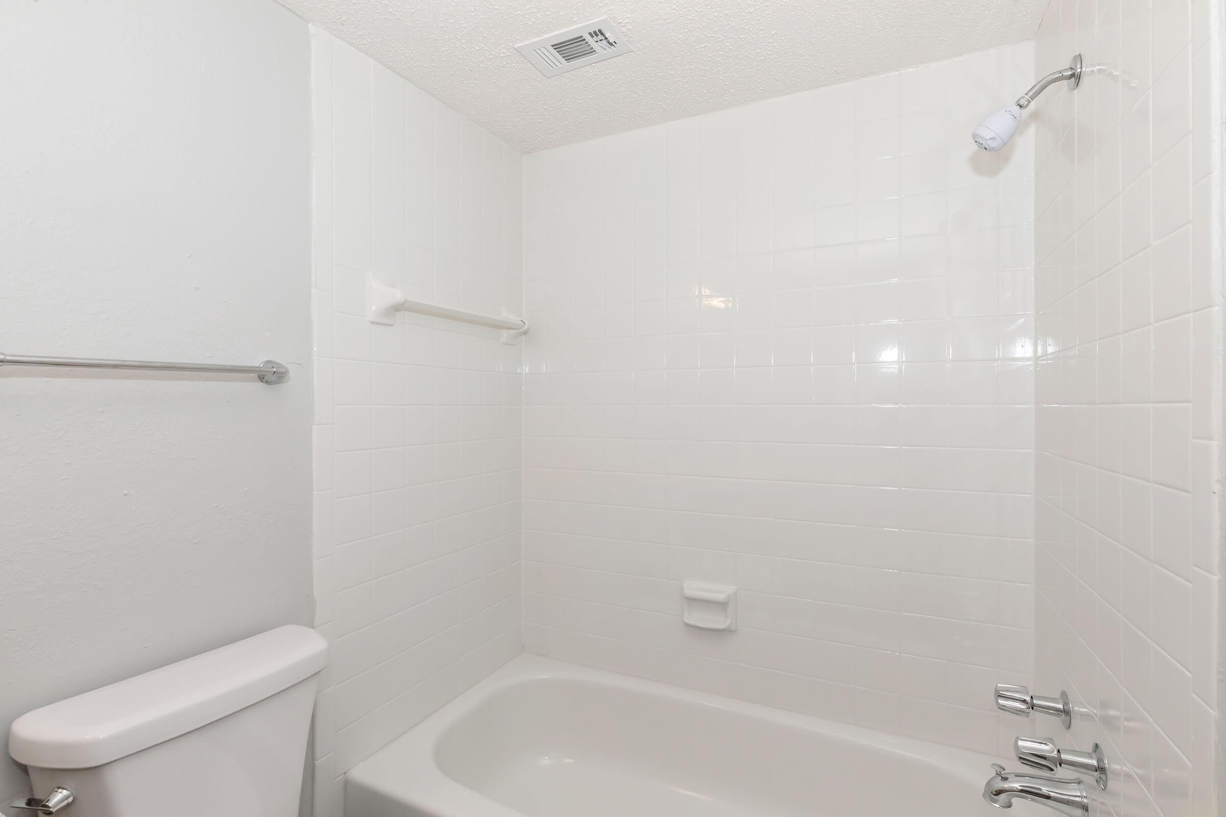 A clean, white-tiled bathroom featuring a bathtub and a showerhead. The wall is mostly white with a towel bar and a soap holder. The toilet is visible on the left side, contributing to a simple and uncluttered design. The overall atmosphere is bright and minimalistic.