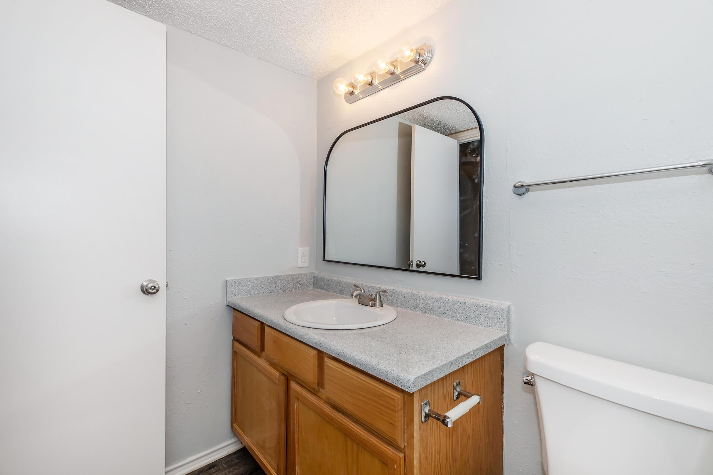 A well-lit bathroom featuring a single sink with a gray countertop, wooden cabinetry, and a large mirror above the sink. There's a white toilet to the right and a towel rack on the wall. The room has a light gray color scheme and a door partially closed, creating a clean and tidy appearance.