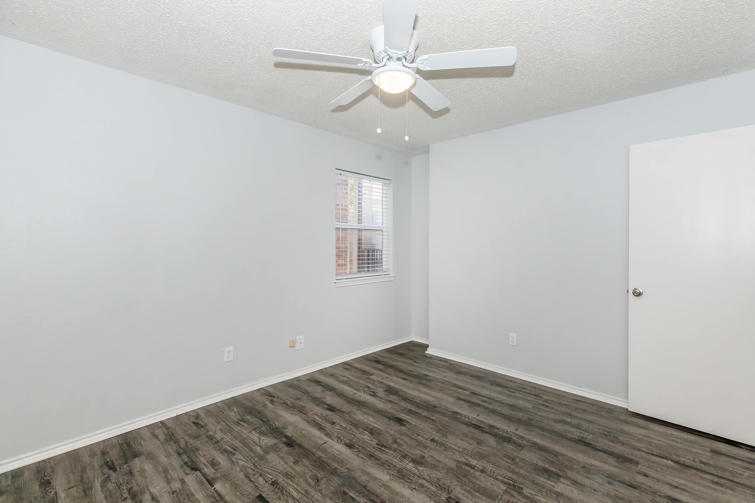 A clean and minimalistic room with light gray walls and a ceiling fan. The floor features wood-like laminate, and there is a window with blinds allowing natural light. A white door is visible on the right side, indicating access to another area. The space appears empty and ready for furniture.