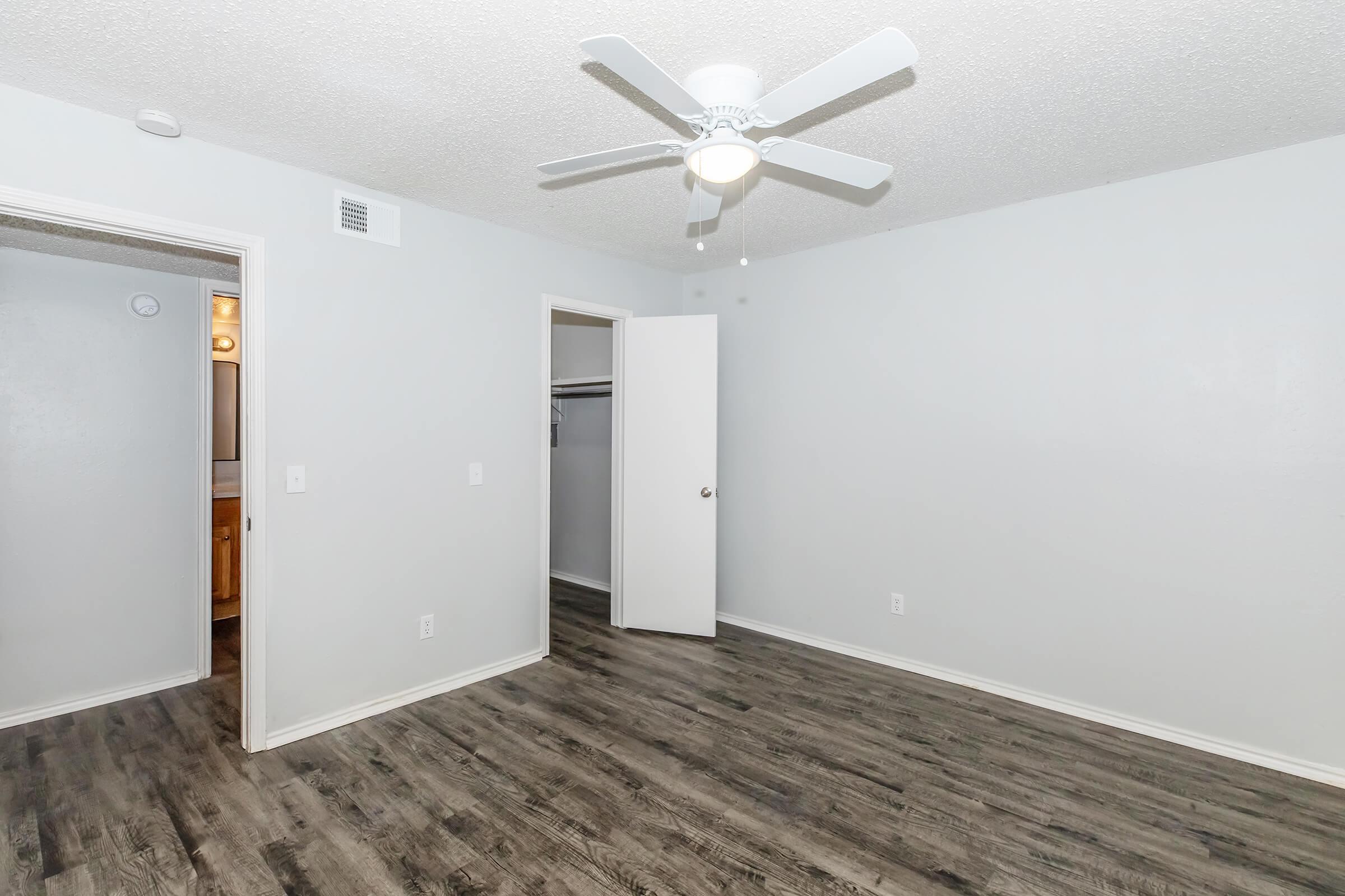 A clean and empty room with light gray walls, a ceiling fan, and dark wood-like flooring. A closet door is slightly open on the left, leading to a bathroom area, while the entrance door is on the right. The space is well-lit and designed for versatility.