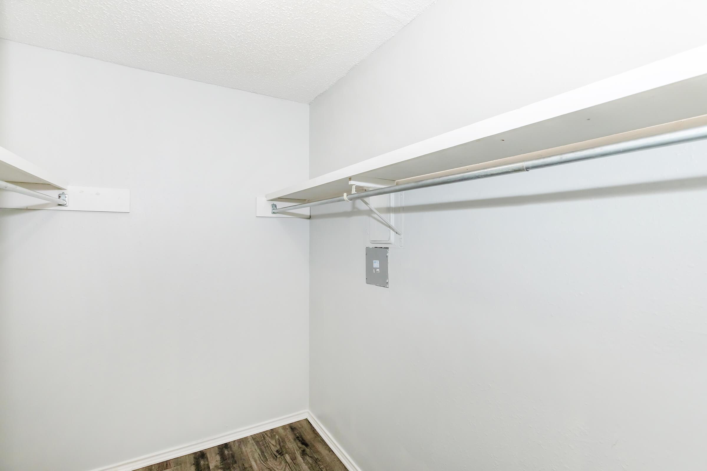 Empty closet with white walls and a wooden floor. The closet features two white shelves running along the top, providing storage space. There are no clothes or items visible, creating a clean, minimalist appearance. A small electrical panel is mounted on the wall.