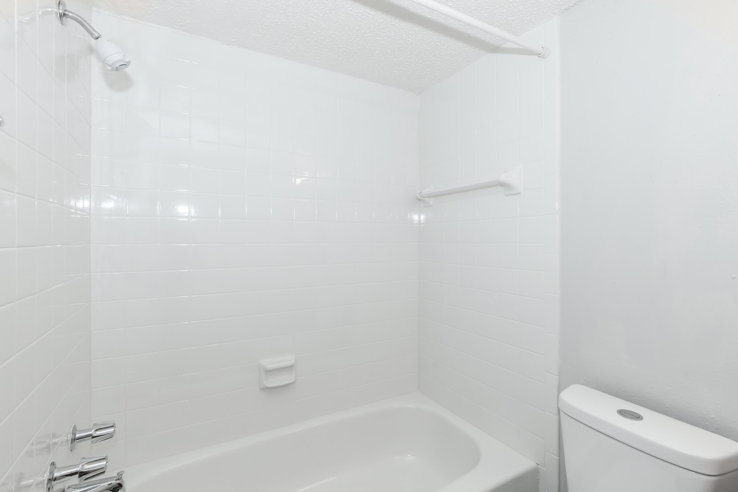 A clean, modern bathroom featuring a white tiled wall, a bathtub with a curved shower rod above it, and a toilet. The space is well-lit and minimalistic, with an emphasis on a fresh, bright aesthetic.