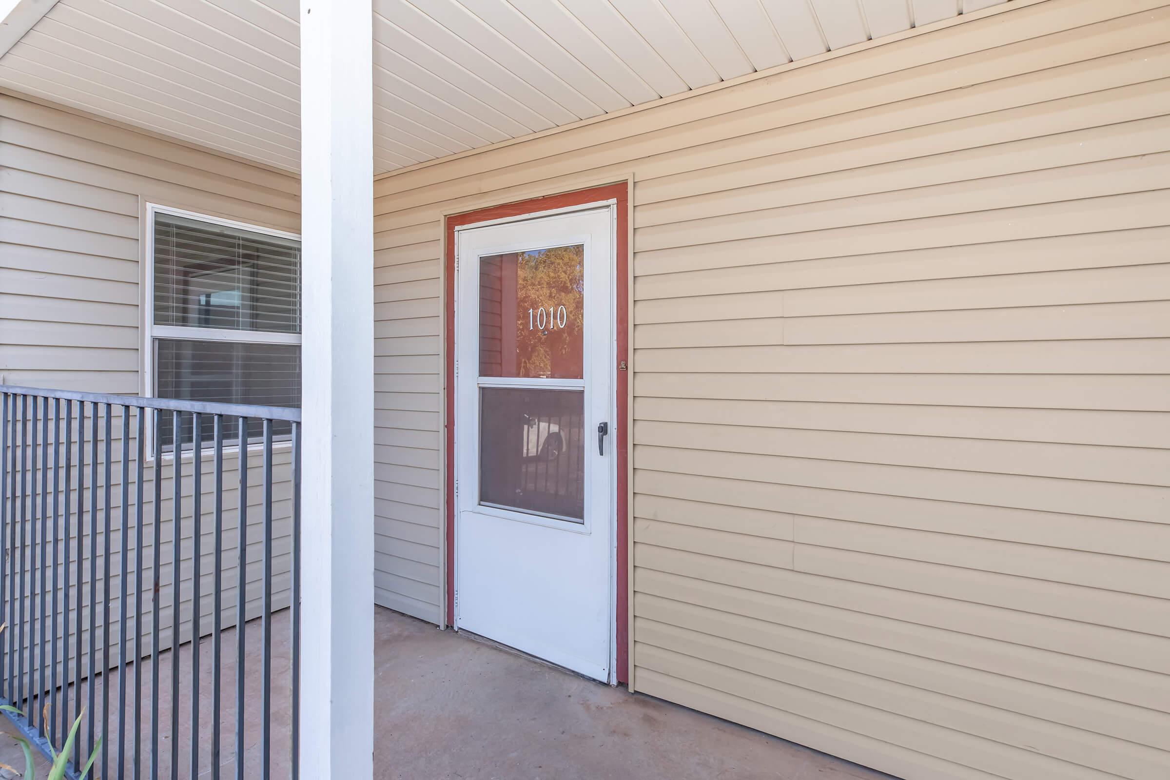 A front porch featuring a white door with a window, marked with the number "1010." The entrance is framed by light-colored siding and a metal railing, with a clear view of the surrounding area. The porch is well-lit and inviting, suggesting a residential setting.