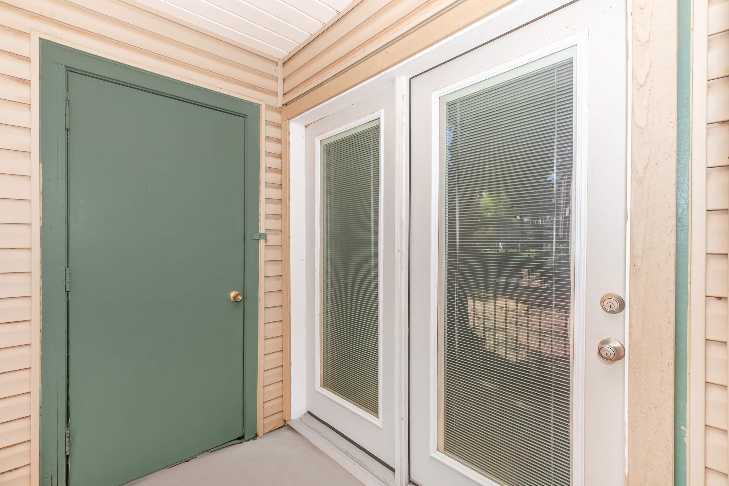 An exterior entrance featuring a green door and double glass doors with vertical blinds. The walls are beige with horizontal siding, and the area is well-lit, showing a glimpse of the outdoor environment through the glass doors.