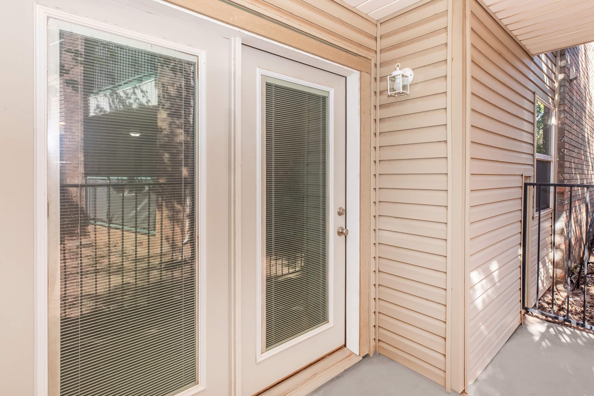 A modern exterior view of a double door with glass panels, framed in white. The door is located on a beige wooden siding wall, featuring a small exterior light fixture to the side. The area has a balcony visible in the background, surrounded by trees and natural light.