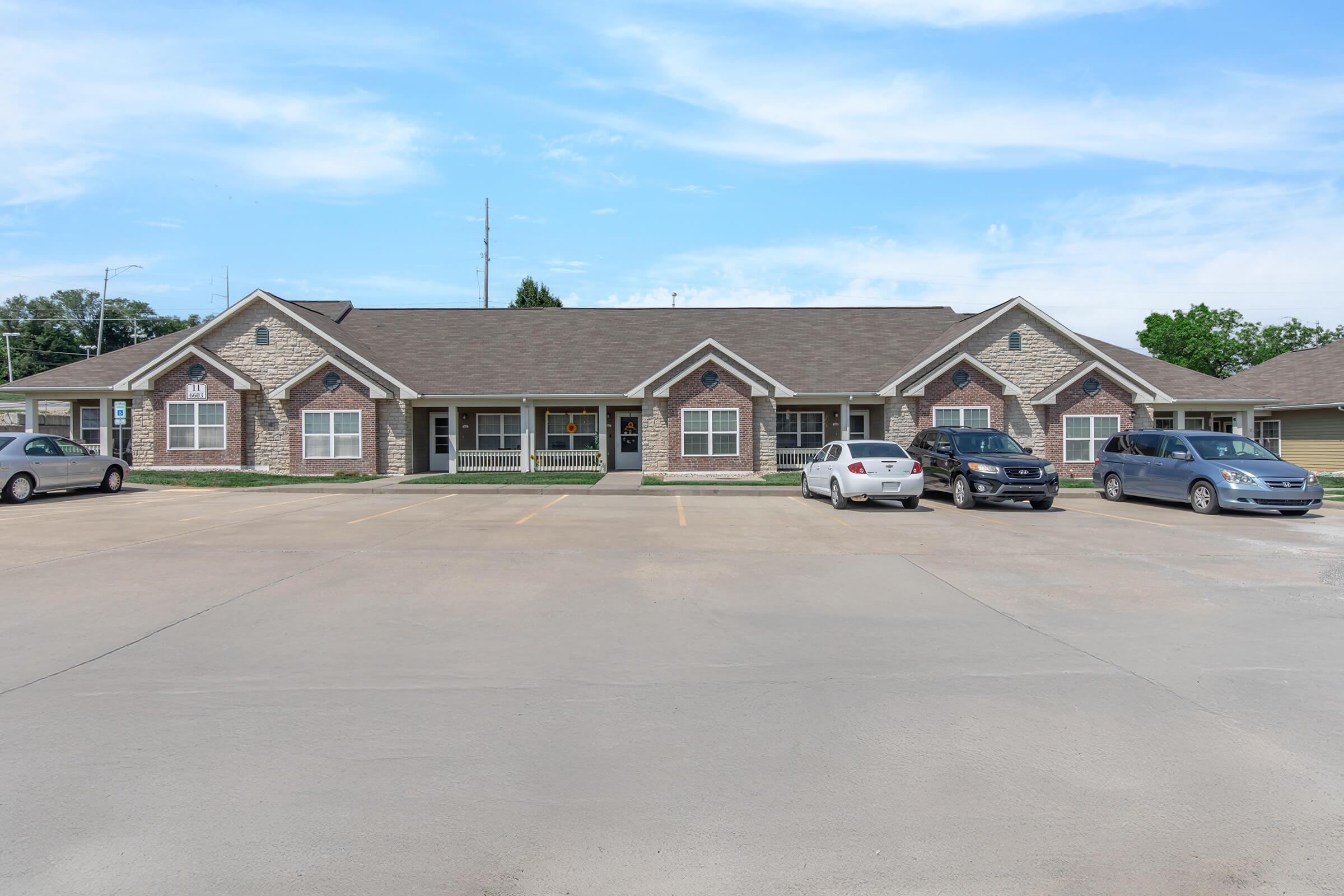 A small commercial building with a brick exterior and multiple sections, featuring large windows and a well-maintained parking lot. Several parked cars are visible in front, and the sky is partly cloudy. Surrounding greenery adds to the pleasant appearance of the area.