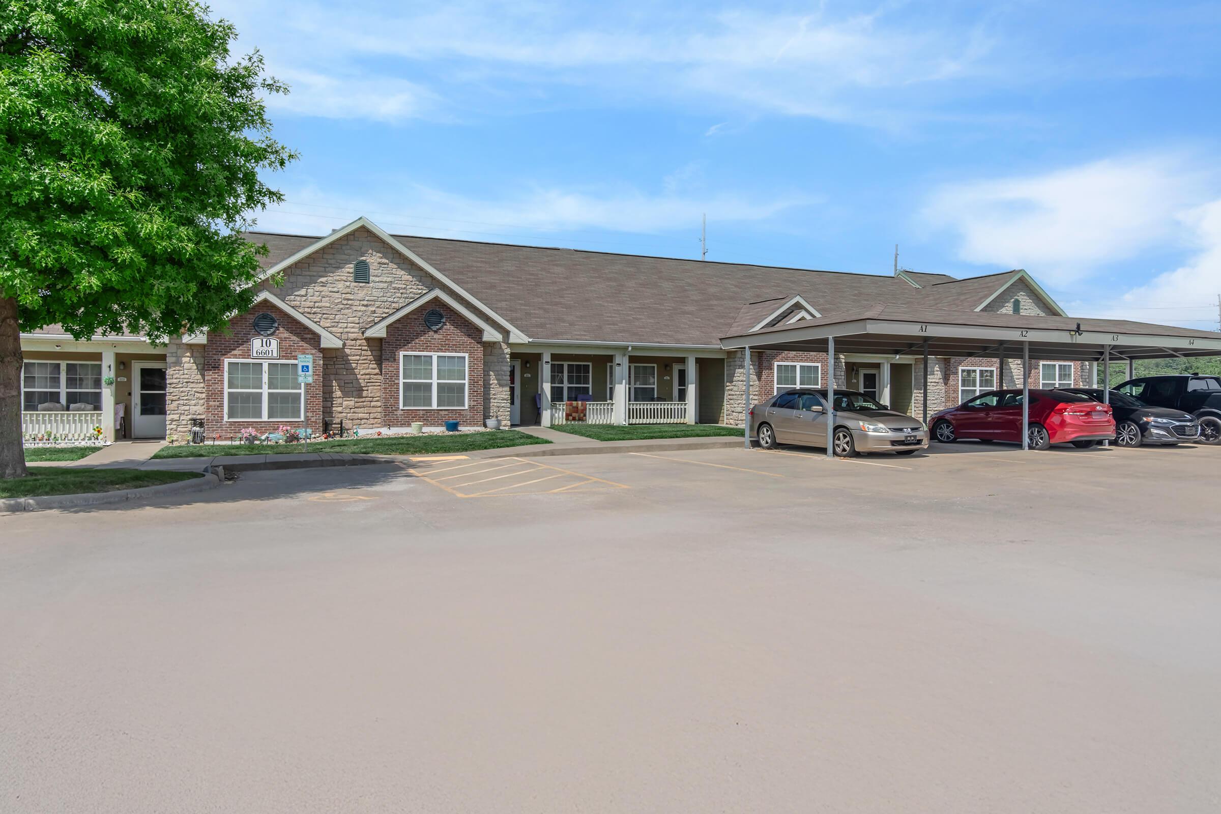 A single-story residential building with a brick facade and a pitched roof. The structure features multiple entrances, a paved parking lot with several parked cars, and trees providing shade in the foreground. The sky is clear with a few clouds.