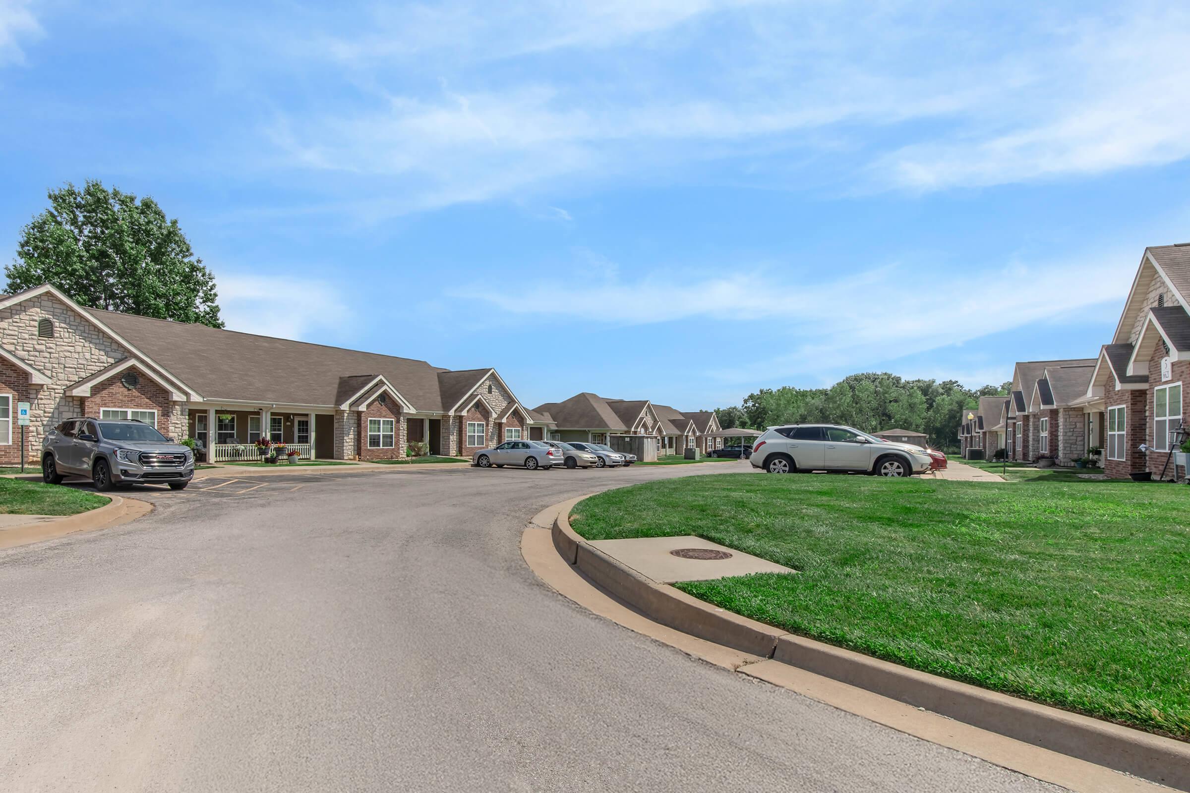 Residential neighborhood with multiple single-story brick homes lining a curved street. Some parked cars are visible along the roadside, and there is a grassy area nearby. The sky is clear with a few clouds, suggesting a sunny day.