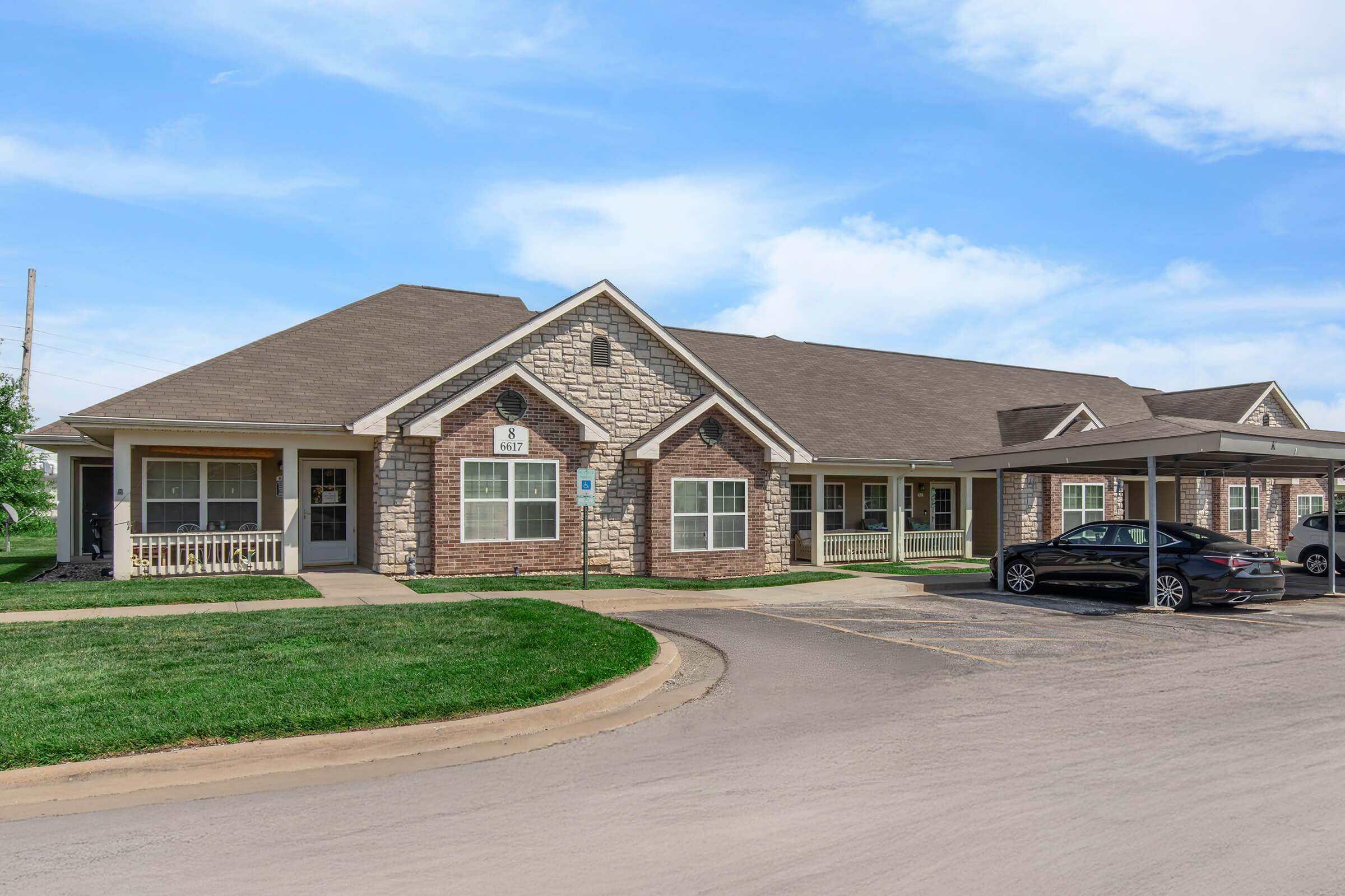A single-story residential building with a brick and stone exterior. The building features multiple windows and a welcoming porch. In the foreground, there's a curved driveway with parked cars. Green grass surrounds the property, set against a clear blue sky.