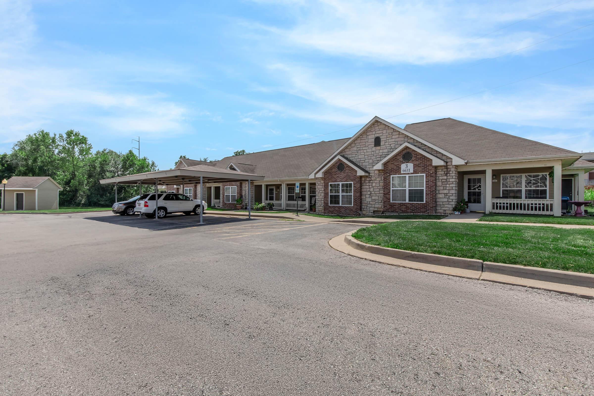 A single-story brick building with a sloped roof, featuring several windows and a porch. In the foreground, there is a paved parking area with vehicles. Surrounding the building are green lawns and trees under a clear blue sky. Additional structures are visible in the background.