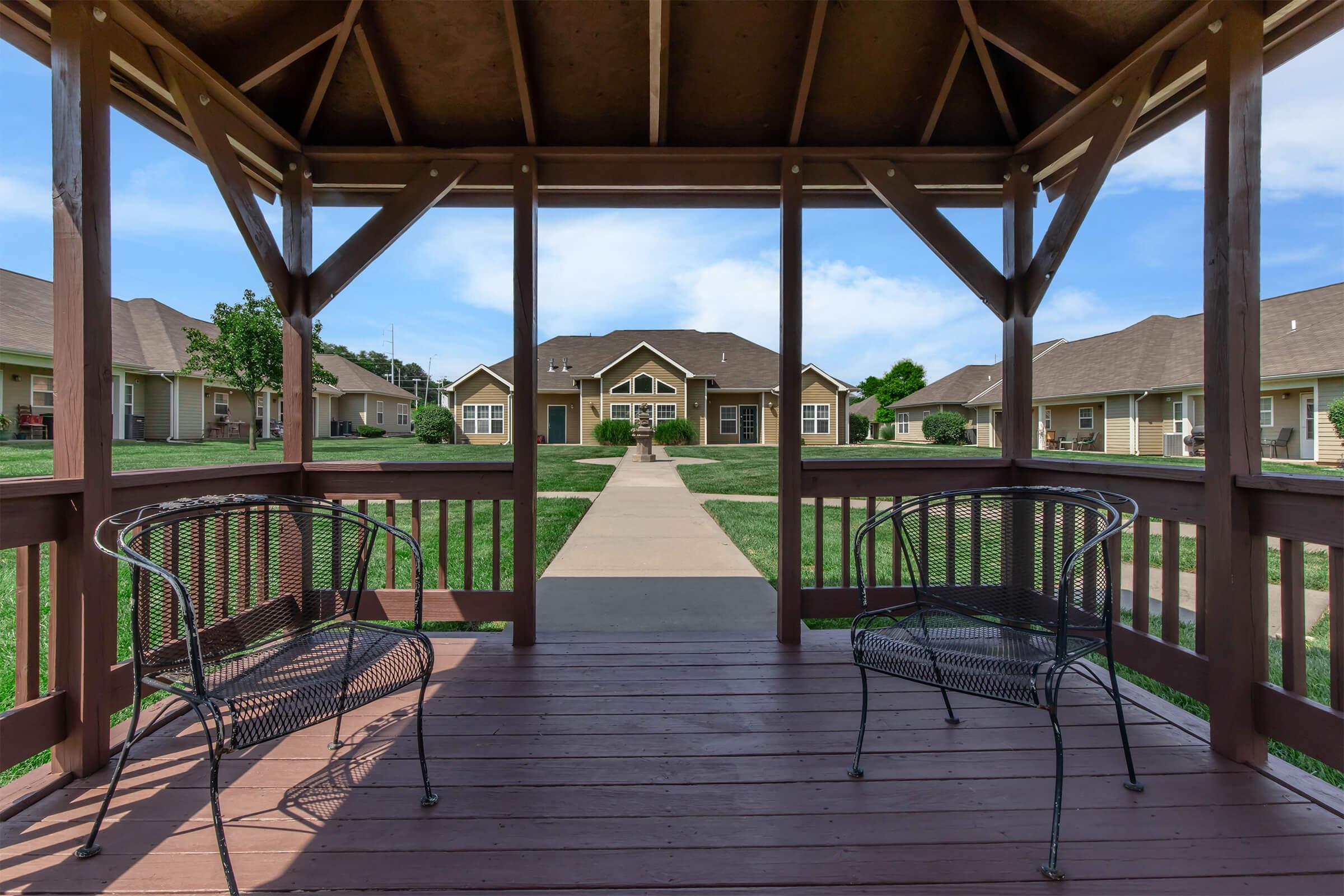 View from a gazebo featuring two empty metal chairs on a wooden deck, overlooking a pathway and a row of houses. The scene is set in a green area with blue skies and fluffy clouds.