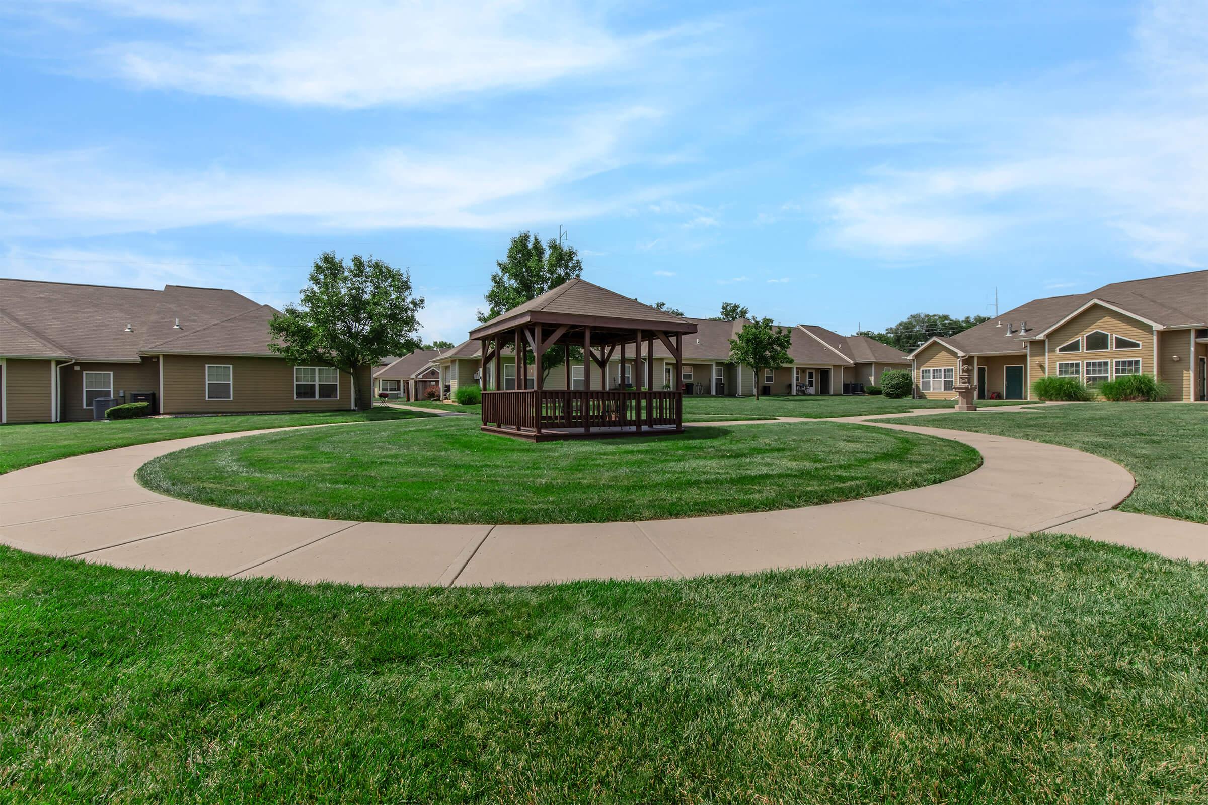A circular pathway surrounds a grassy area with a gazebo in the center. The background features several modest homes with beige siding and brown roofs, under a blue sky with light clouds. The scene conveys a peaceful, residential atmosphere.