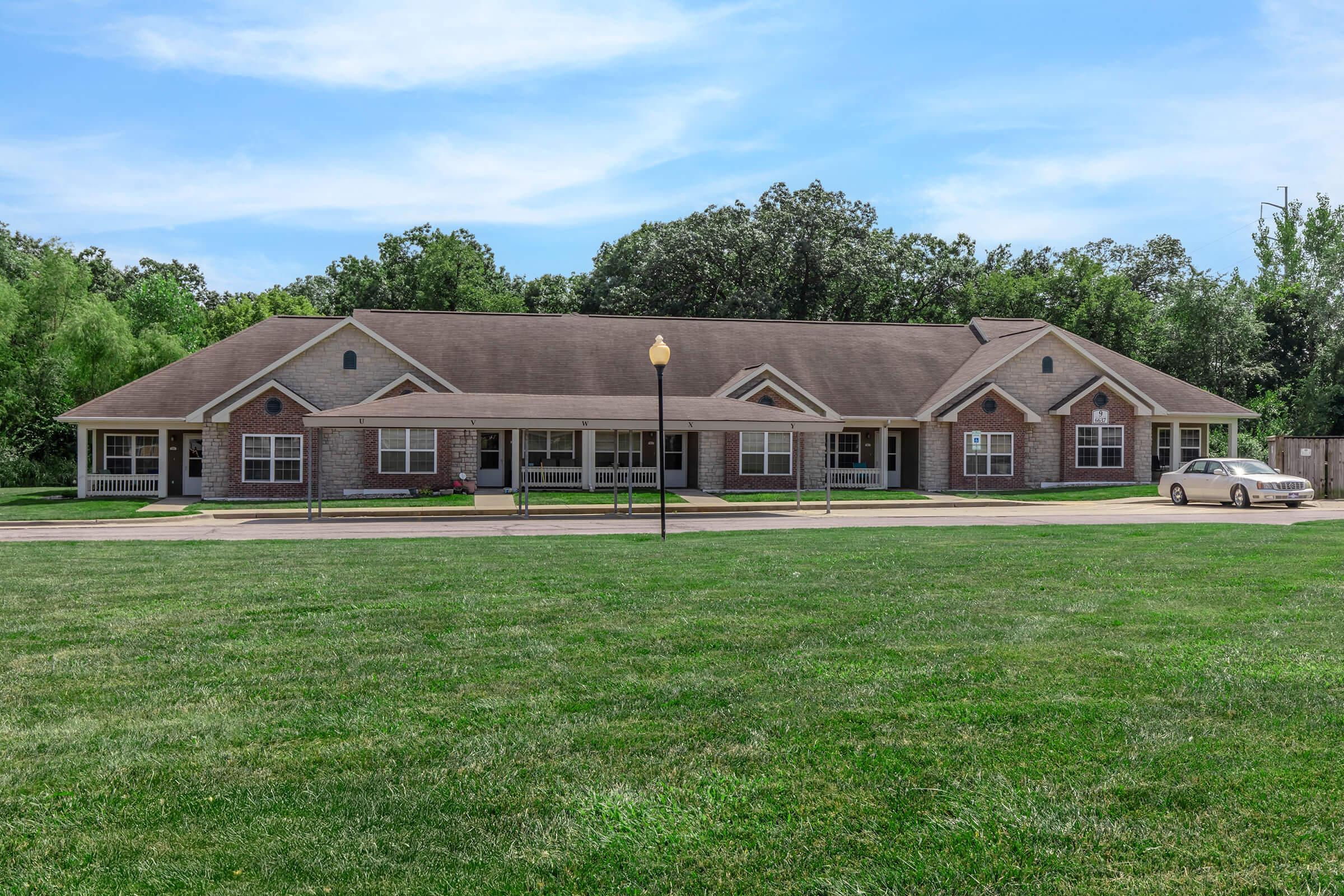 A single-story brick building with multiple windows and a pitched roof, surrounded by a well-manicured grassy area. There are trees in the background and a light pole in front. A white car is parked on the driveway in front of the building. The sky is clear with a few clouds.