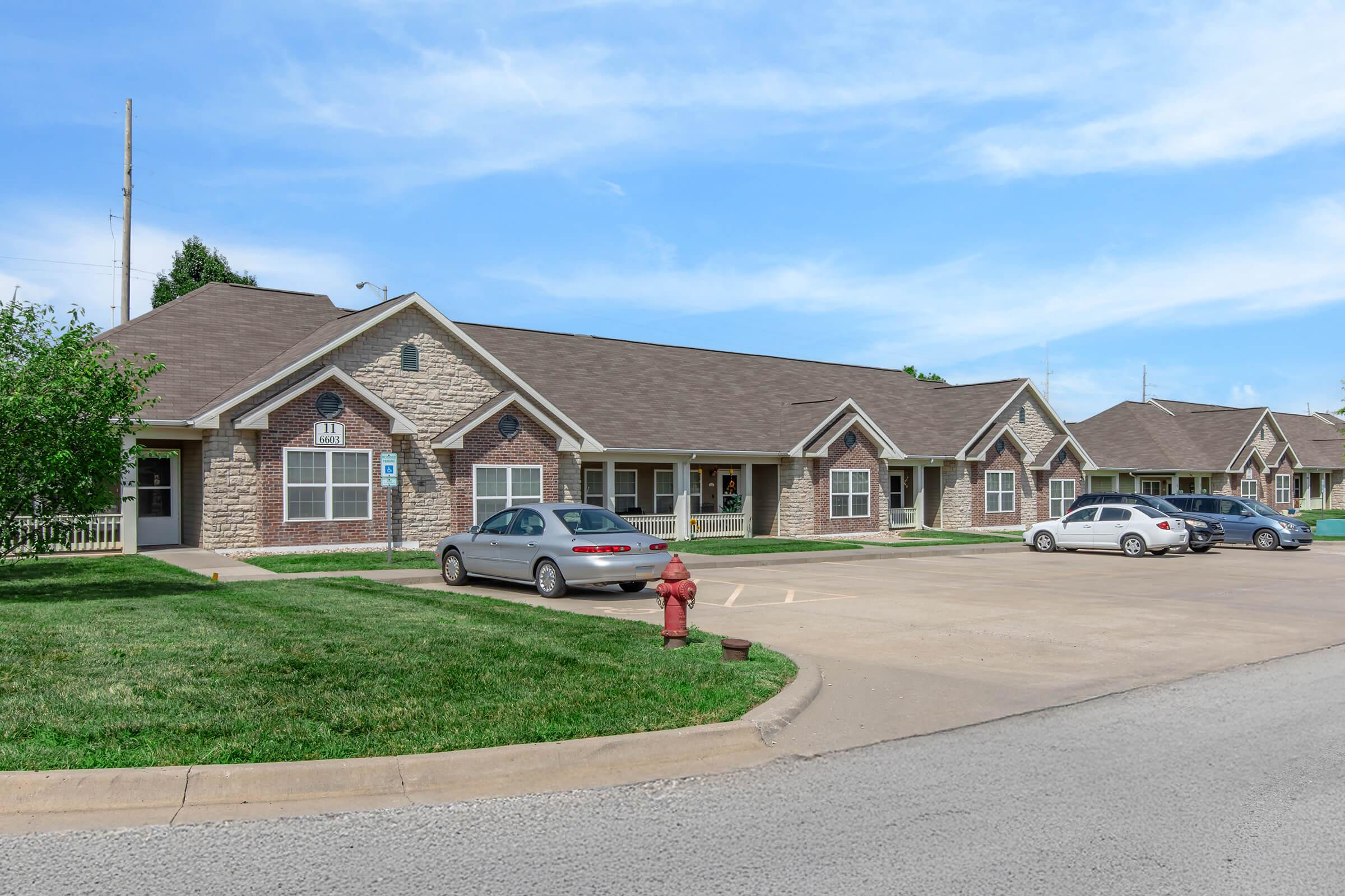 A row of single-story brick and siding buildings with a well-maintained lawn in front. There are parked cars in the lot and a red fire hydrant beside the driveway. The sky is blue with a few clouds, indicating a clear day.