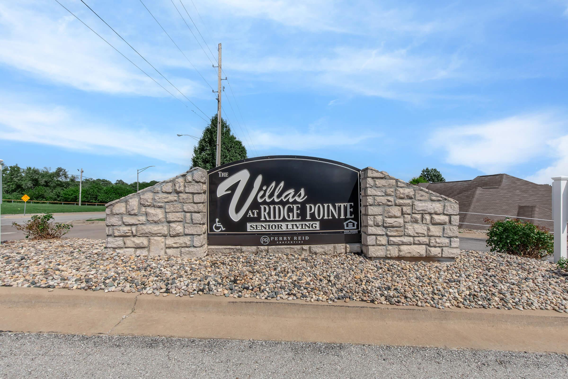 Sign for "The Villas at Ridge Pointe Senior Living," featuring a black background with white text, set against a stone base. The area is landscaped with gravel and grass, and the sky is clear with a few clouds. Nearby infrastructure indicates a community setting.