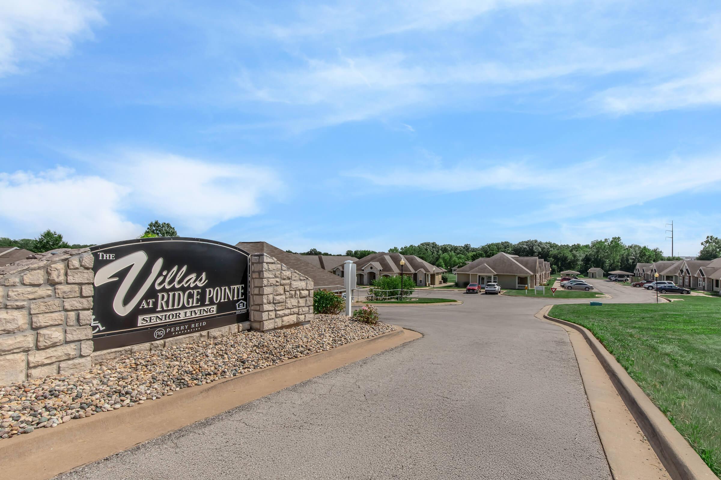 Sign for "The Villas at Ridge Pointe," a senior living community, featuring a stone border. A paved road leads into the complex, with residential buildings visible in the background under a clear blue sky. Lush green grass is on the sides of the road.