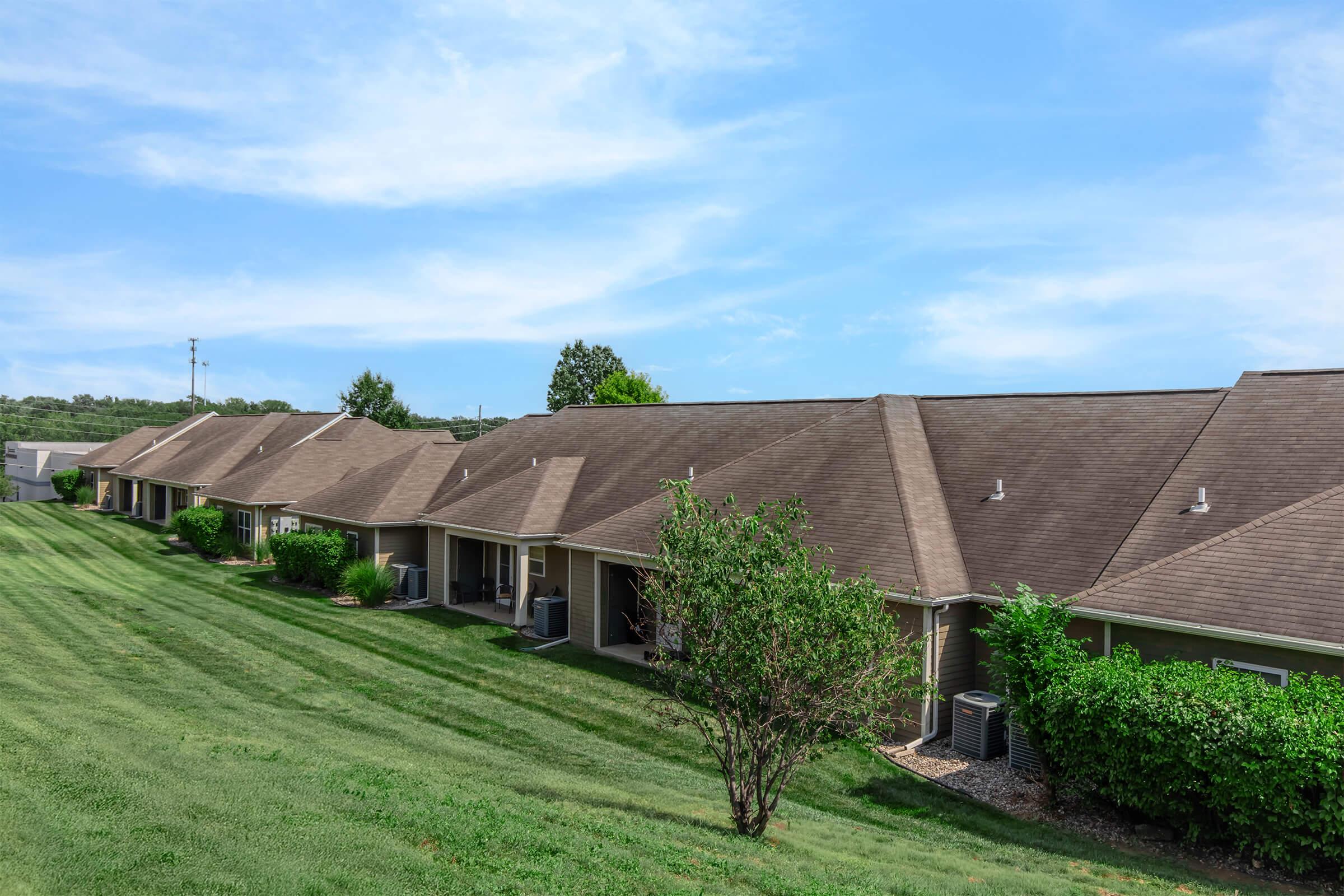 A row of single-story houses with brown roofs, set on a well-maintained green lawn. The sky is bright blue with a few clouds. Some trees are visible in the background, and air conditioning units are placed by the sides of the houses. The scene conveys a peaceful suburban atmosphere.