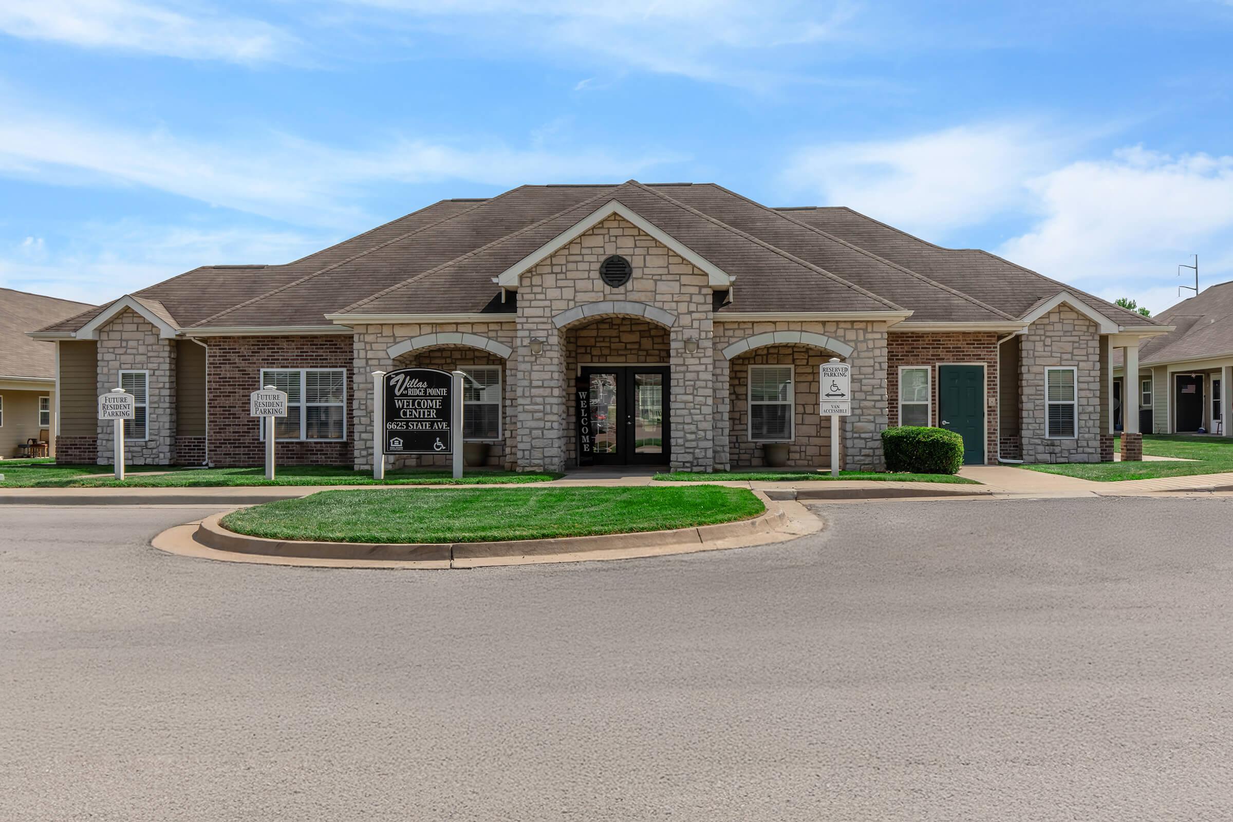 A single-story stone and brick building with a sloped roof, featuring large front windows and double doors. There are signs in front, and well-maintained green lawns on either side. The building is situated on a paved road with a clear blue sky above.