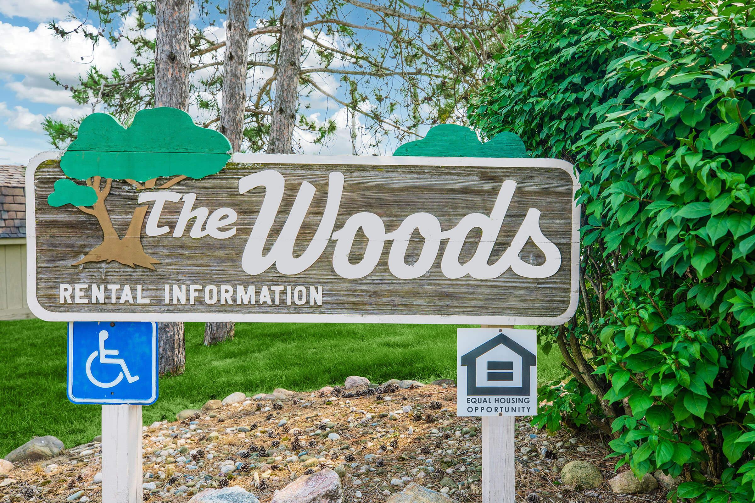 Sign for "The Woods" featuring rental information, with a wooden design and decorative trees. The area includes a blue wheelchair accessibility symbol and a symbol promoting equal housing opportunity. The background shows green grass and shrubbery under a partly cloudy sky.