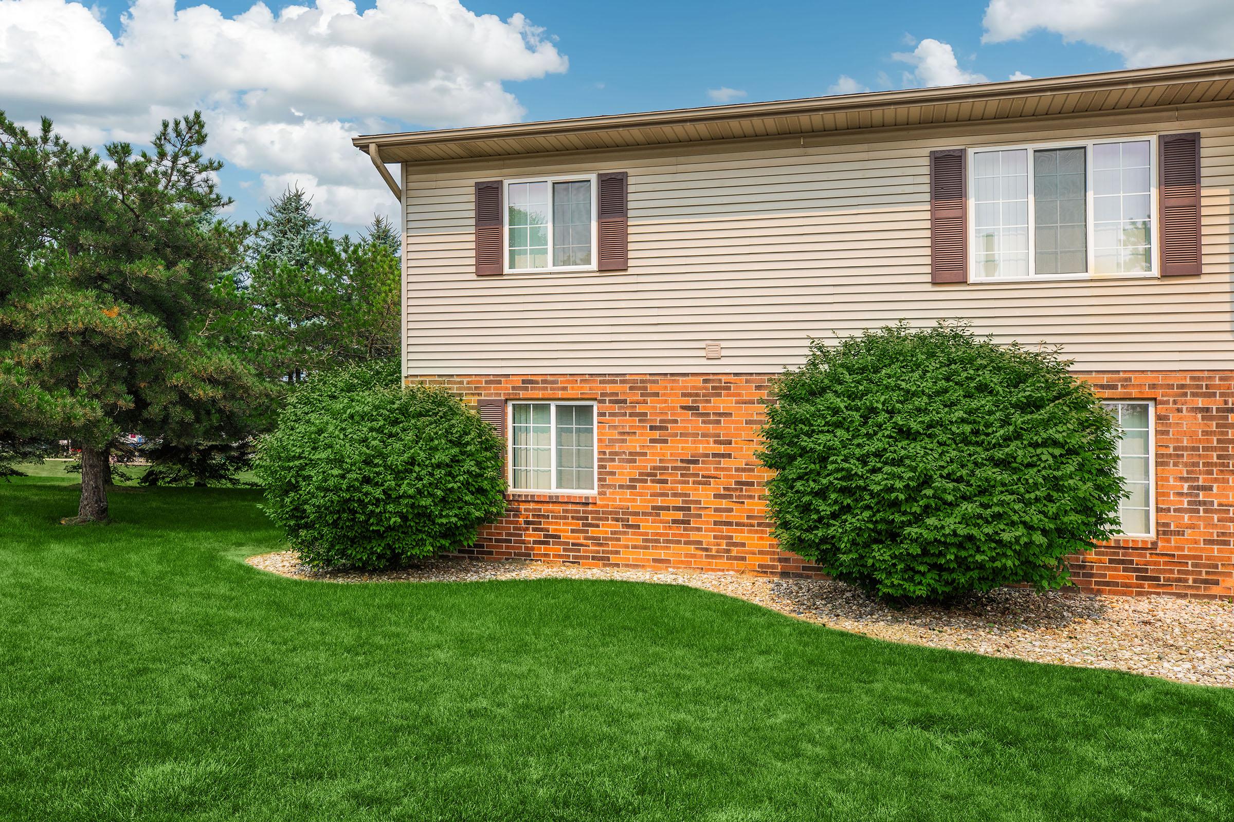A well-maintained exterior view of a two-story building with beige siding and dark brown shutters. Two rounded green shrubs are positioned near the foundation, and there's a vibrant green lawn surrounding the building. In the background, trees and a partly cloudy sky are visible.