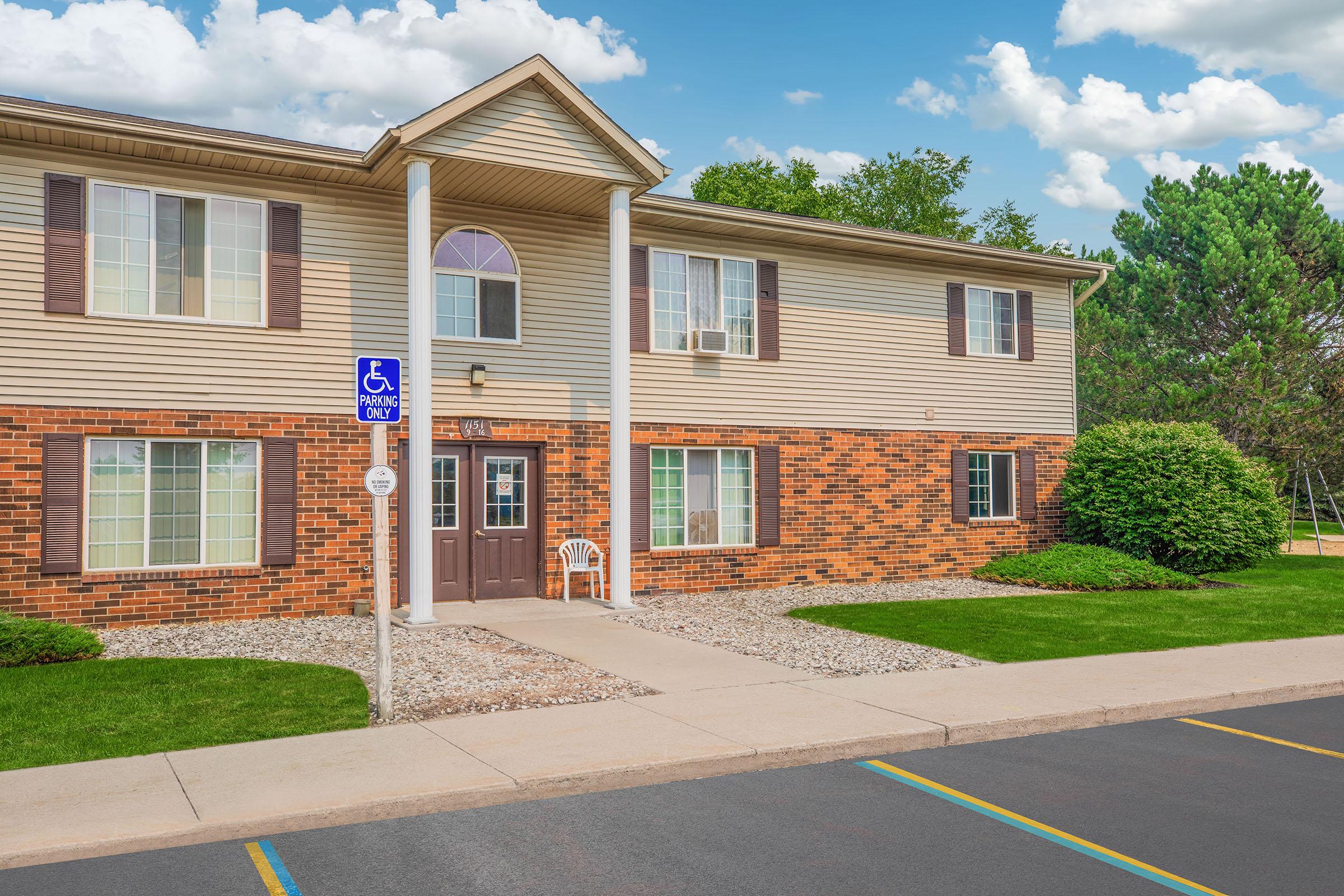 A two-story residential building with a beige facade and red brick accents. The entrance features a covered porch with a chair, flanked by windows. A handicapped parking sign is visible, and well-maintained green grass surrounds the building, with trees and a playground in the background under a partly cloudy sky.