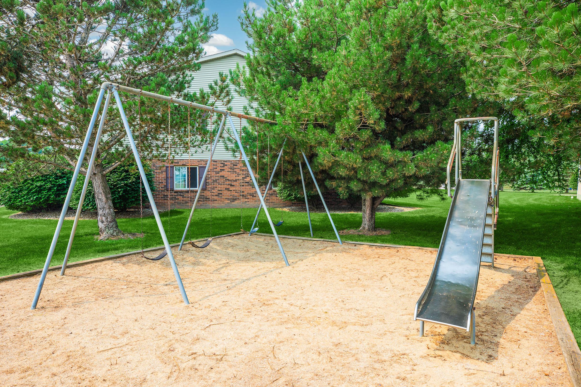 A playground featuring two swings and a metal slide, set on a sandy surface. Surrounding the area are green grass and several trees, with a brick building visible in the background under a clear blue sky.