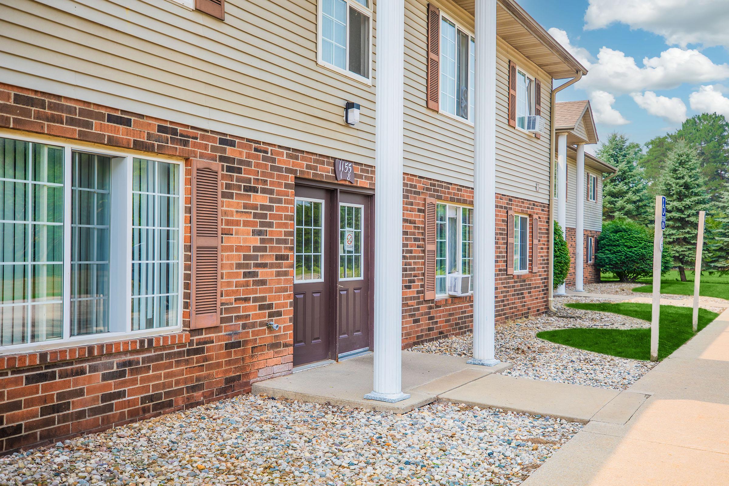 Exterior view of a two-story building with brick and siding. The entrance features double doors framed by white columns. Large windows with divided panes line the front, and the pathway is bordered by pebbles and grass. Lush greenery is visible in the background under a partly cloudy sky.